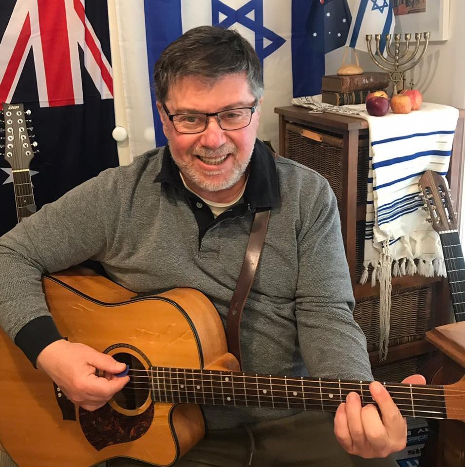 A smiling man wearing glasses and a gray sweater playing an acoustic guitar indoors. Behind him are flags, a menorah, and fruit on a table.