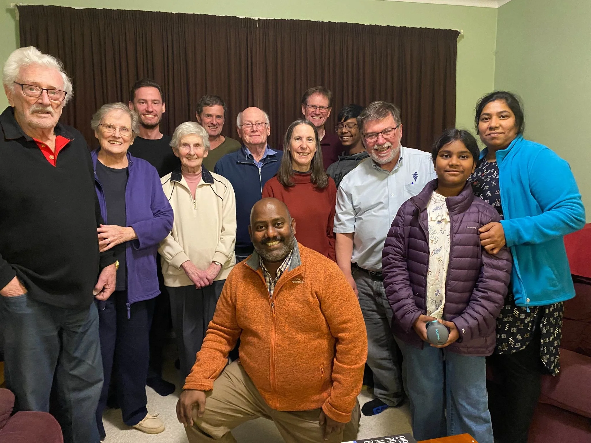 A diverse group of 14 people gathered indoors, smiling for a group photo. They are standing in front of a brown curtain, with some seated in front. The individuals vary in age, gender, and ethnicity, wearing casual and winter clothing.