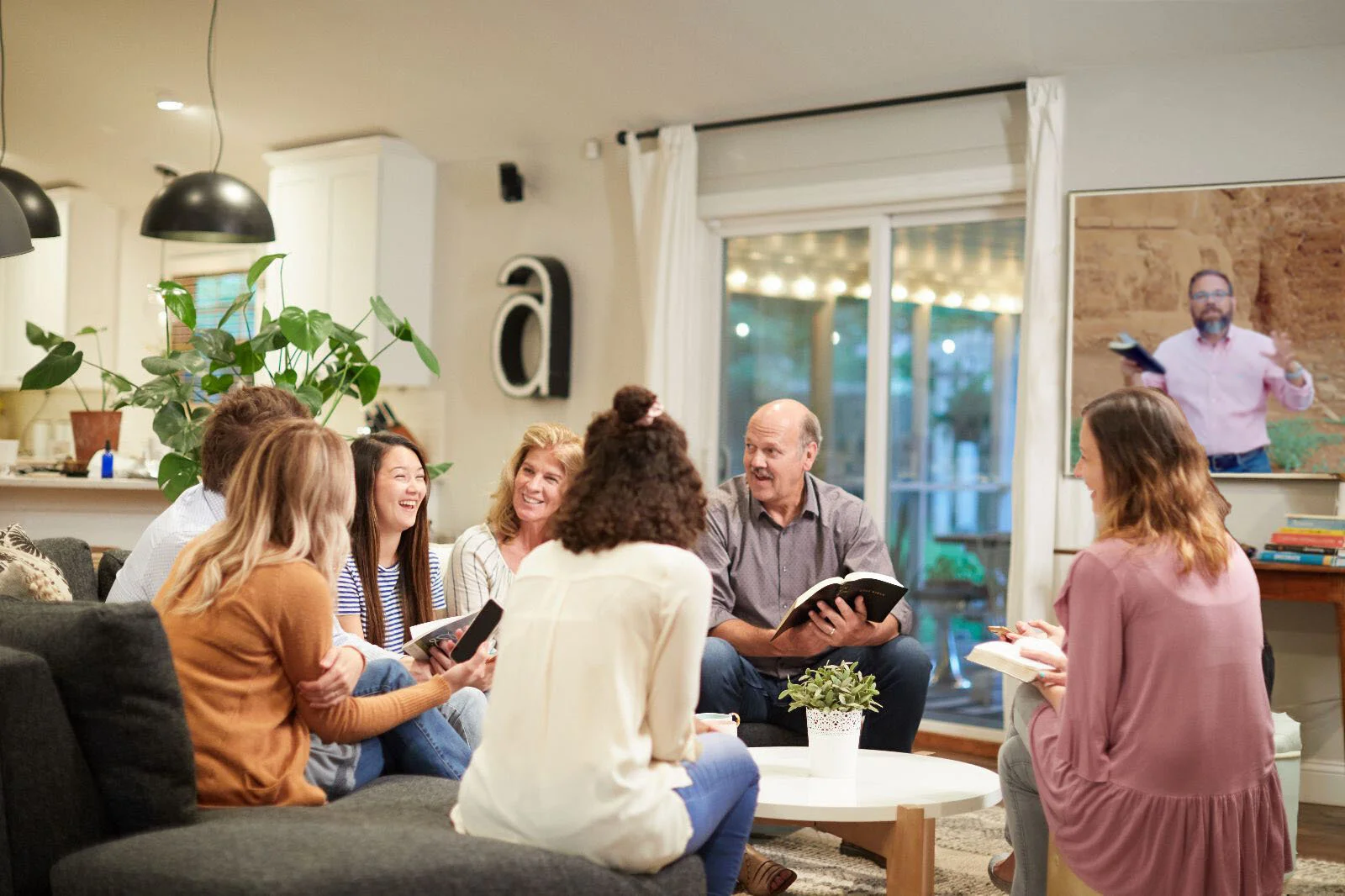 Group of people sitting in a living room, engaged in a discussion or group activity, with a man holding a book and a woman taking notes.