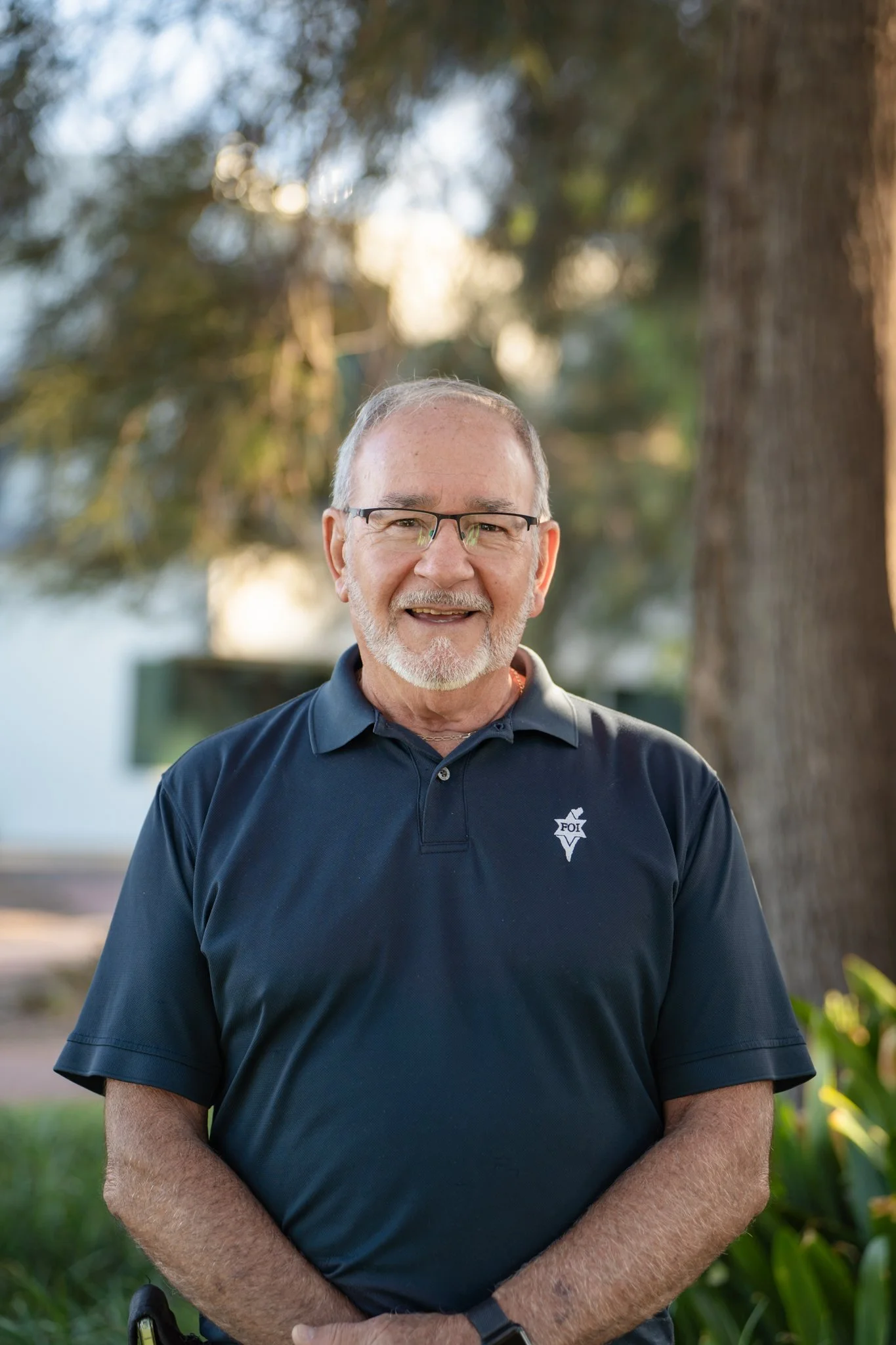 A smiling man with glasses and a beard wearing a collared shirt, standing indoors near a window with a brick wall background.