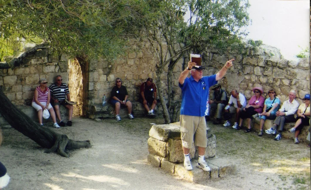 Tour guide pointing and talking to a group of tourists seated outdoors near stone ruins and trees.