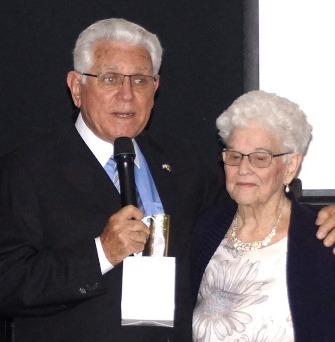 An elderly man in a black suit, holding a microphone, standing next to an elderly woman with white hair, glasses, wearing a necklace and floral blouse, appearing at an indoor event.