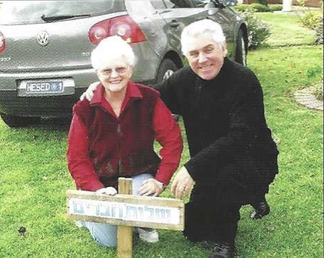 An elderly woman and an elderly man kneeling on the grass in front of a parked gray Volkswagen car, smiling for a photo, with a small wooden sign held by the woman.