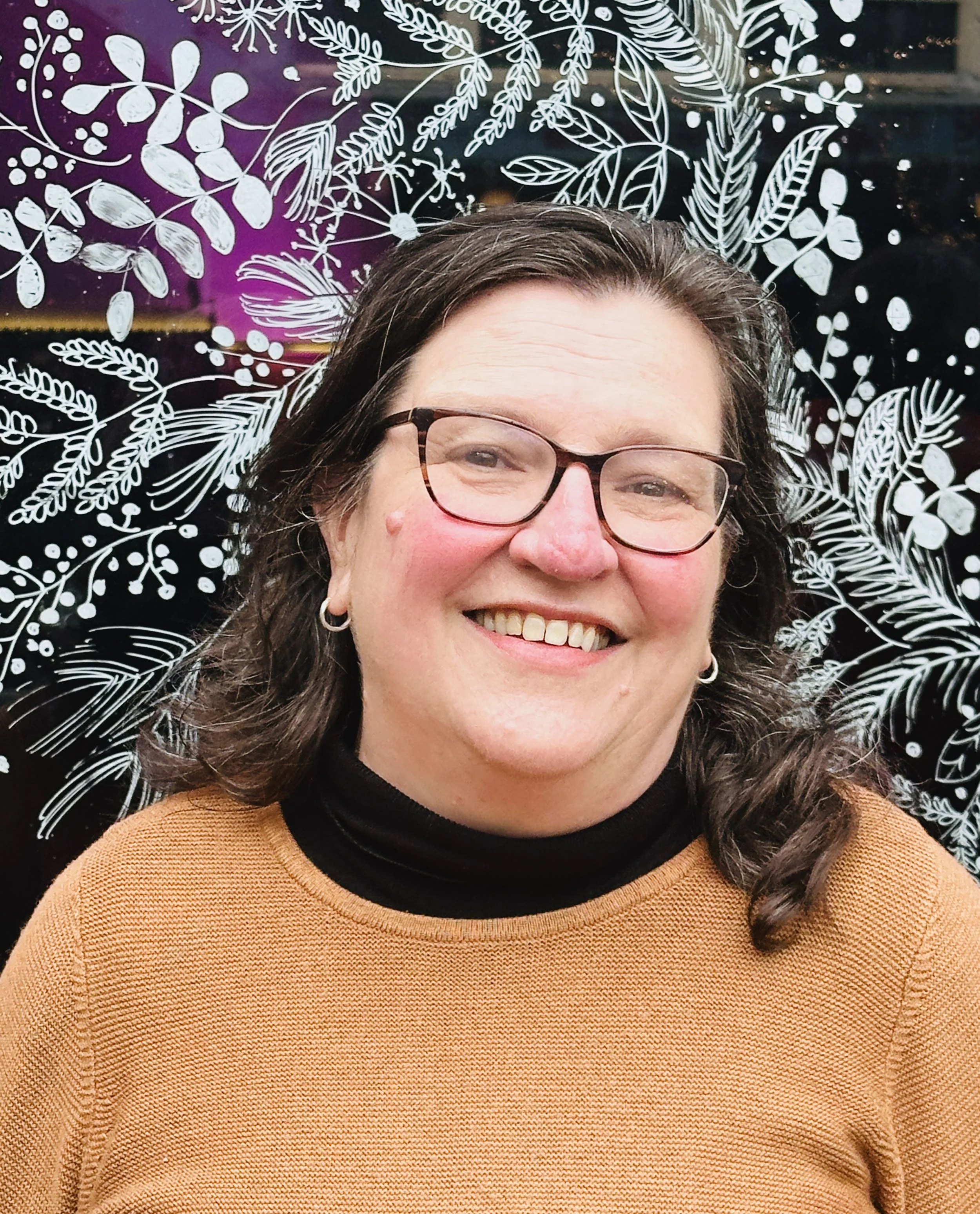 A smiling woman with glasses and short dark hair wearing a red textured sweater with buttons on the shoulder, standing in front of a wooden door.