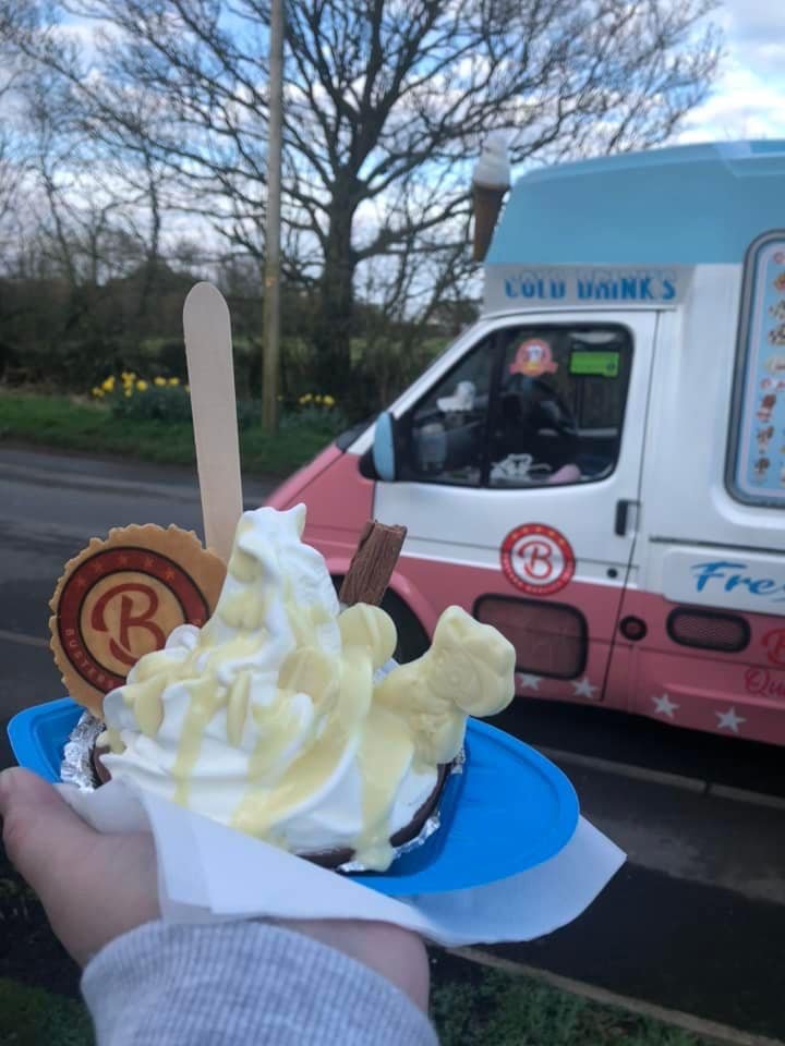 A hand holding a cup of vanilla soft serve ice cream with chocolate and vanilla drizzle, a chocolate stick, a cookie, and a wafer on a blue tray. In the background, there is an ice cream truck with a pink and white design and a tree with no leaves.