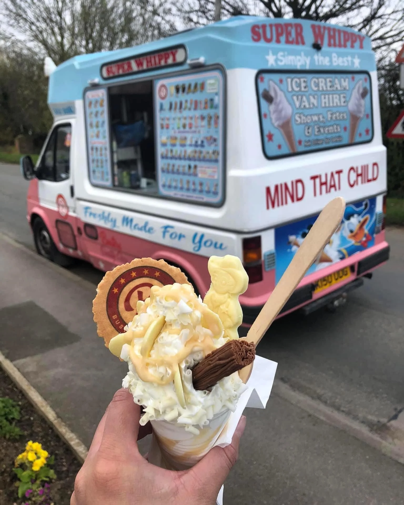 A person holding a vanilla ice cream cone with toppings including whipped cream, chocolate, and a cookie, with a colorful ice cream truck in the background.
