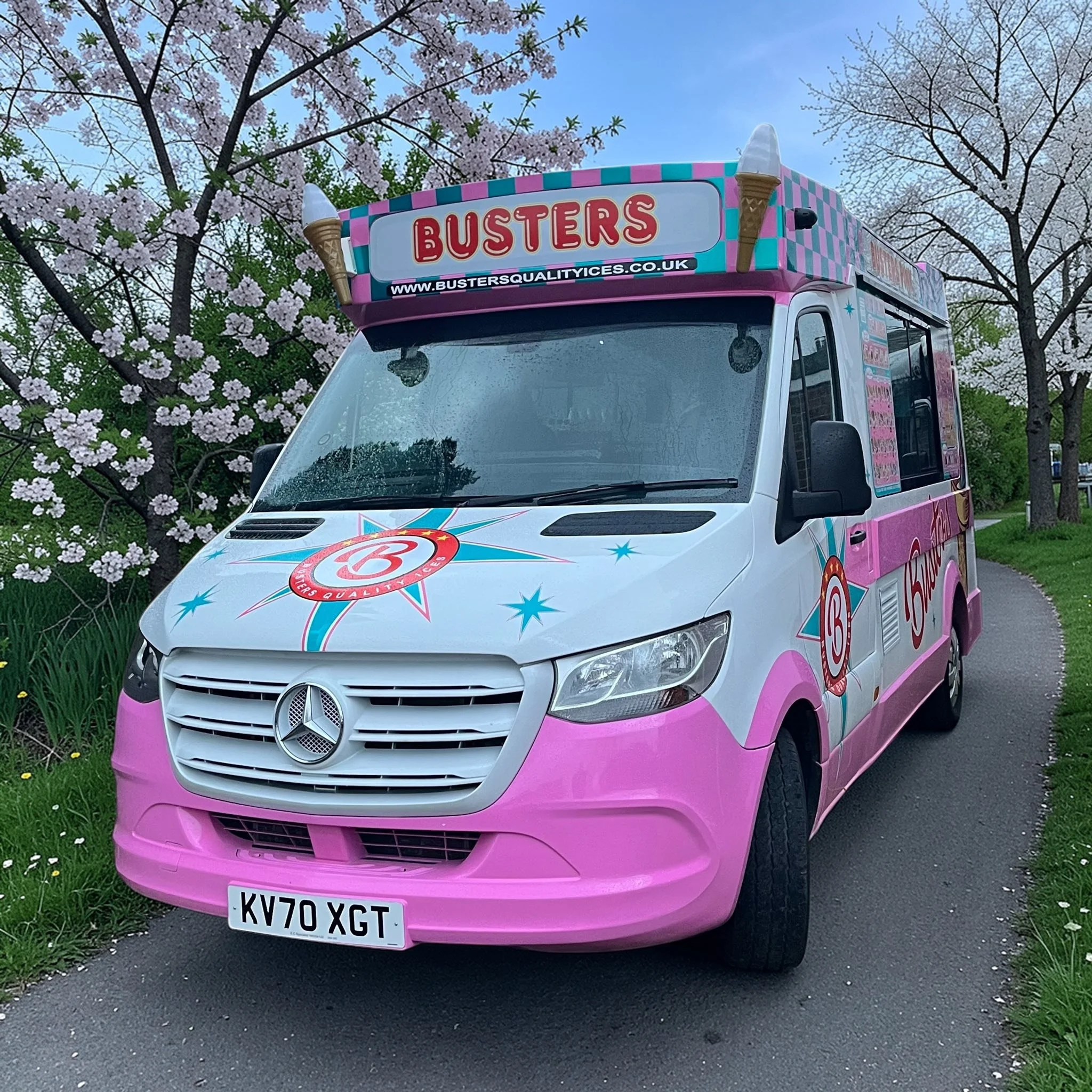 An ice cream truck decorated with pink, blue, and white colors, featuring ice cream cone images and the word "BUSTERS" on the sign. The truck is parked on a street with pink flowering trees and a partly cloudy sky in the background.