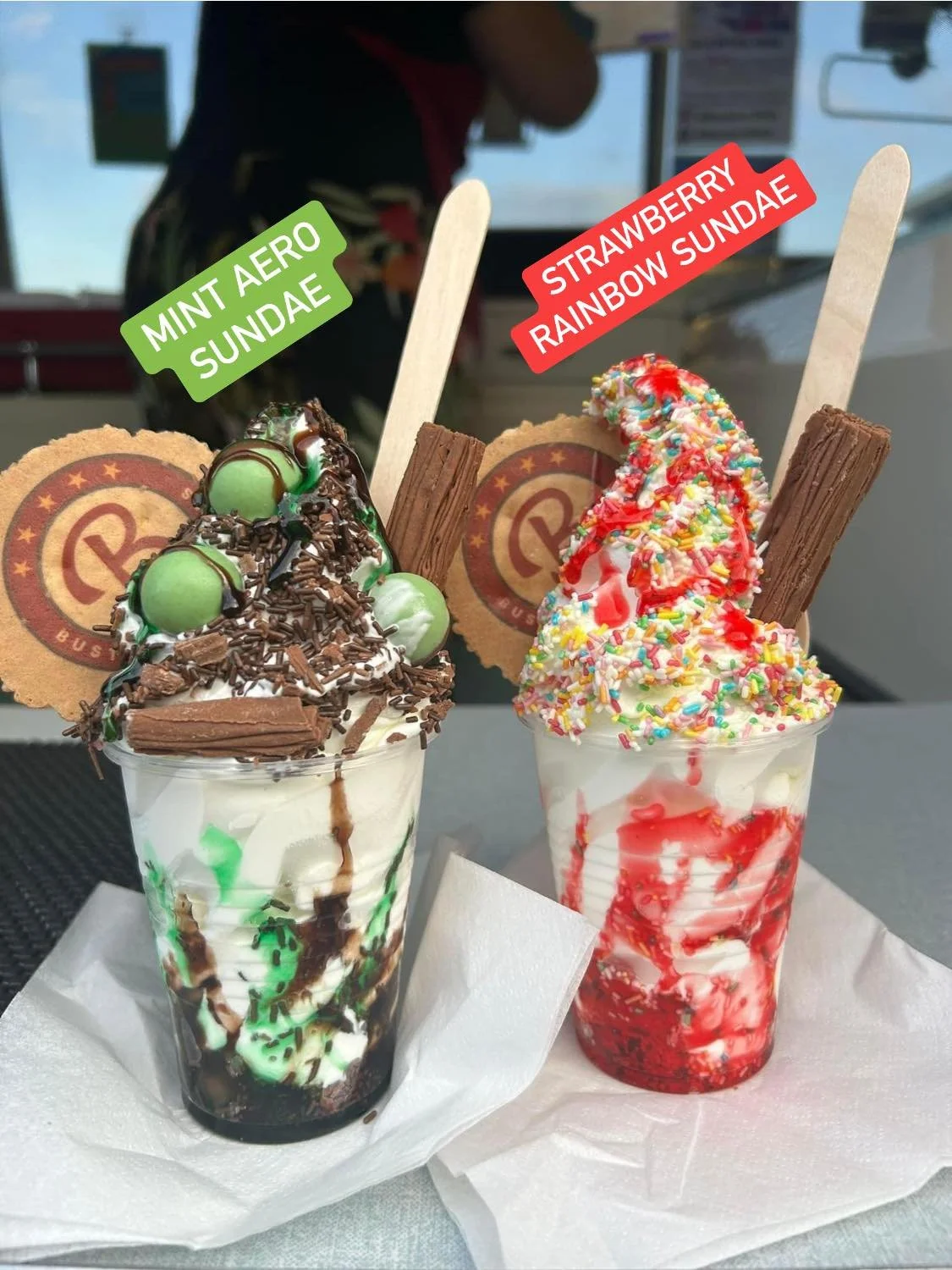 Two ice cream sundaes in cups with toppings and labels. The left sundae is labeled 'Mint Aero Sundae' and has green and chocolate candies, whipped cream, chocolate shavings, and a wafer. The right sundae is labeled 'Strawberry Rainbow Sundae' and has