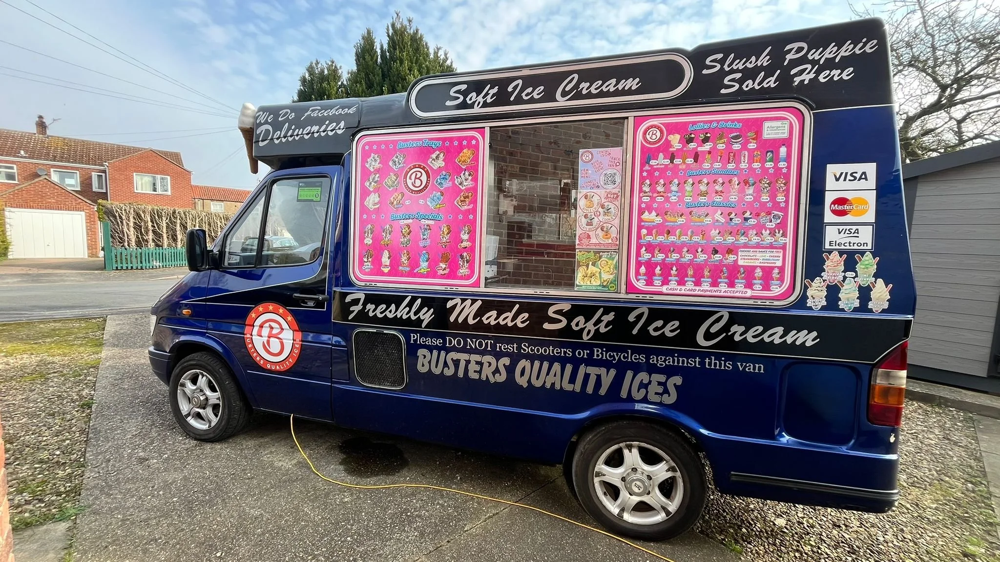 Blue ice cream truck offering various frozen treats, including soft ice cream, slush puppies, and ice cream in toppings trays, parked on a street with houses and trees in the background.