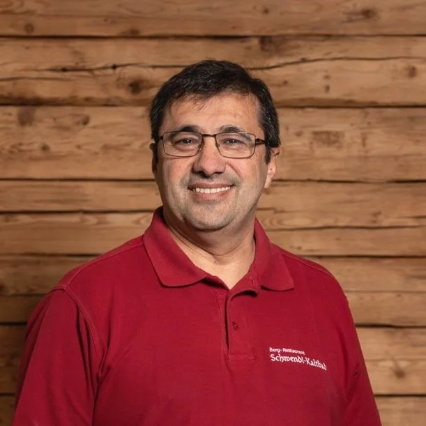 Ein Mann mit Brille trägt ein rotes Polo-Shirt mit der Aufschrift "Berg-Restaurant Schoenb-Kaltbad". Er steht vor einem Hintergrund aus Holzplanken und lächelt in die Kamera.
