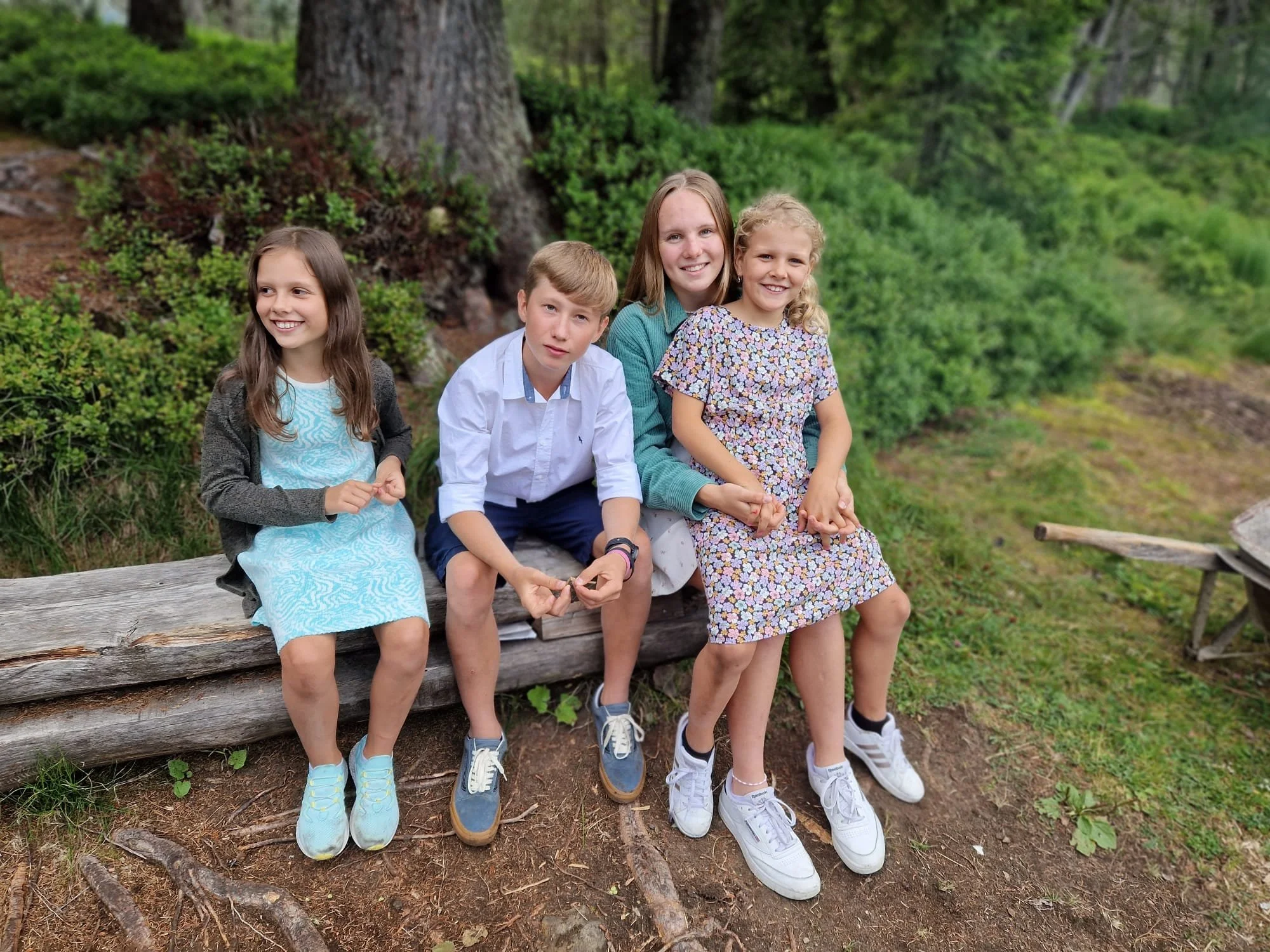 Vier Kinder sitzen auf einer Holzbank und lächeln in einer Waldumgebung. Zwei Mädchen tragen Kleider, eines davon ist blau und das andere blumig. Der Junge trägt ein weißes Hemd. Im Hintergrund sind Bäume und Gebüsch zu sehen.