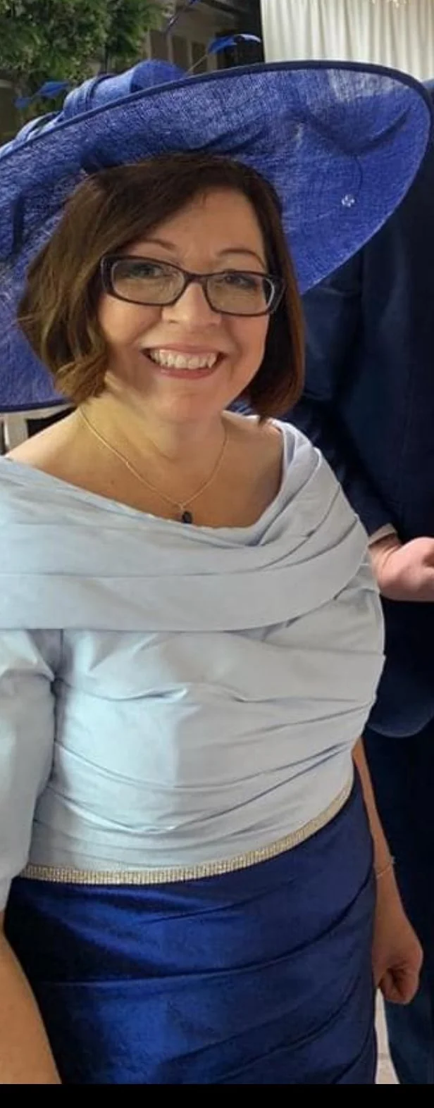 A woman with short brown hair, glasses, and a big smile, wearing a light gray off-the-shoulder top, a dark blue skirt, a gold necklace, and a large blue hat, standing outdoors.