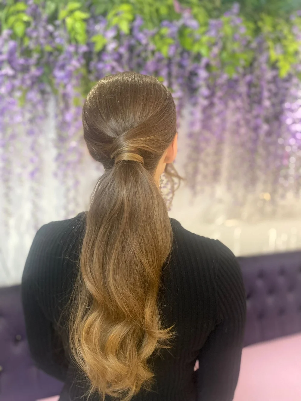 A woman with long, wavy, light brown hair styled in a half-updo, seen from behind, sitting in front of a purple floral background.
