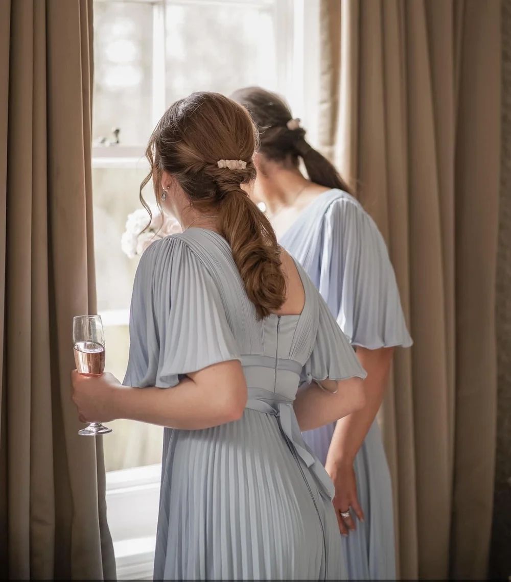 Two women in light gray pleated dresses looking out a window, one holding a glass of pink beverage, with curtains framing the scene.