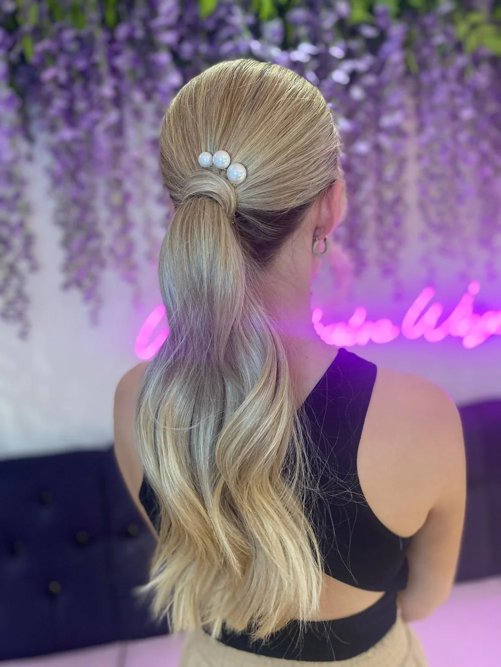 Back view of a woman with long, blonde wavy hair styled in a low ponytail decorated with pearl hairpins, standing in front of a wall adorned with purple flowers and pink neon signage.