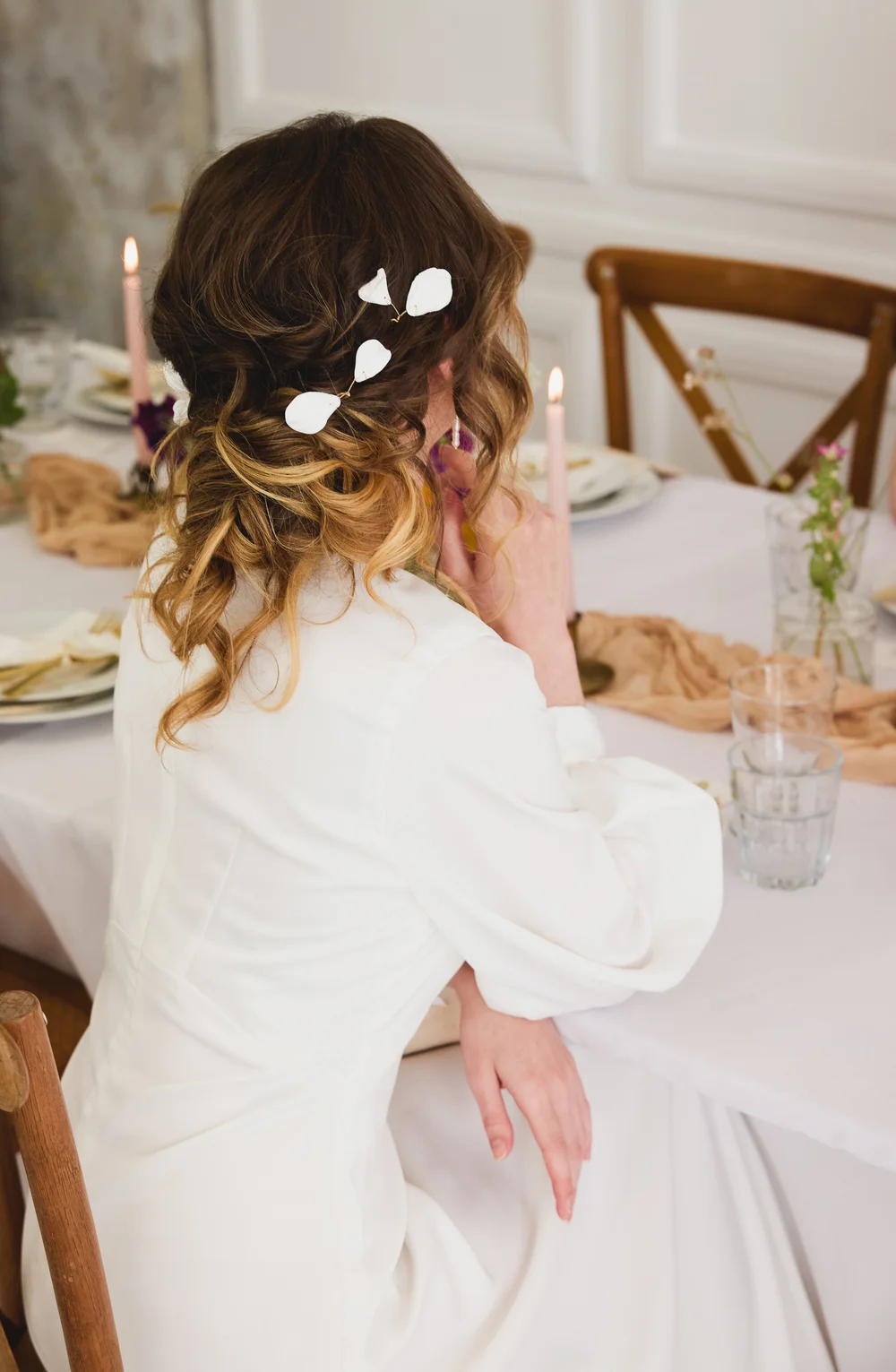 A woman with curly, shoulder-length hair decorated with white leaf-shaped accessories, sitting at a dining table with candles and floral arrangements.