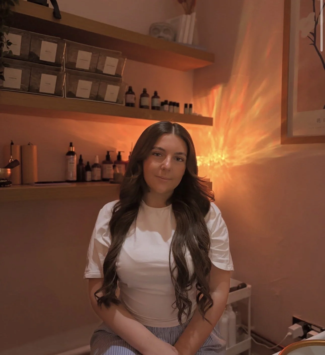 A young woman with long, straight blonde hair and makeup, standing in front of purple hanging wisteria flowers and a pink neon sign that says "Fly". She wears a white t-shirt, black pants, and jewelry, and is smiling softly.