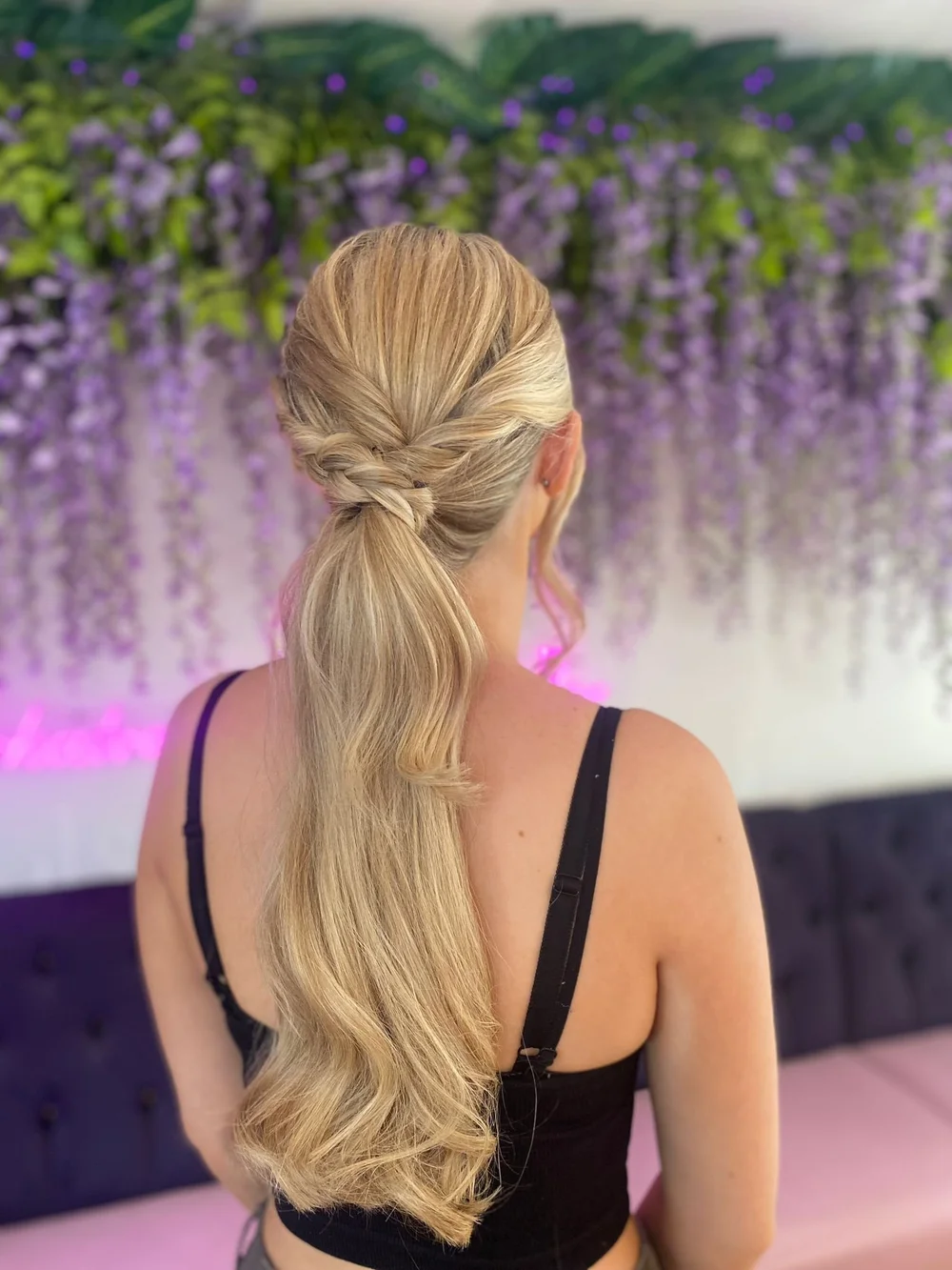 Back of woman with long blonde hair styled in a twisted low ponytail, wearing a black spaghetti strap top, in front of a purple wisteria floral backdrop with green leaves.