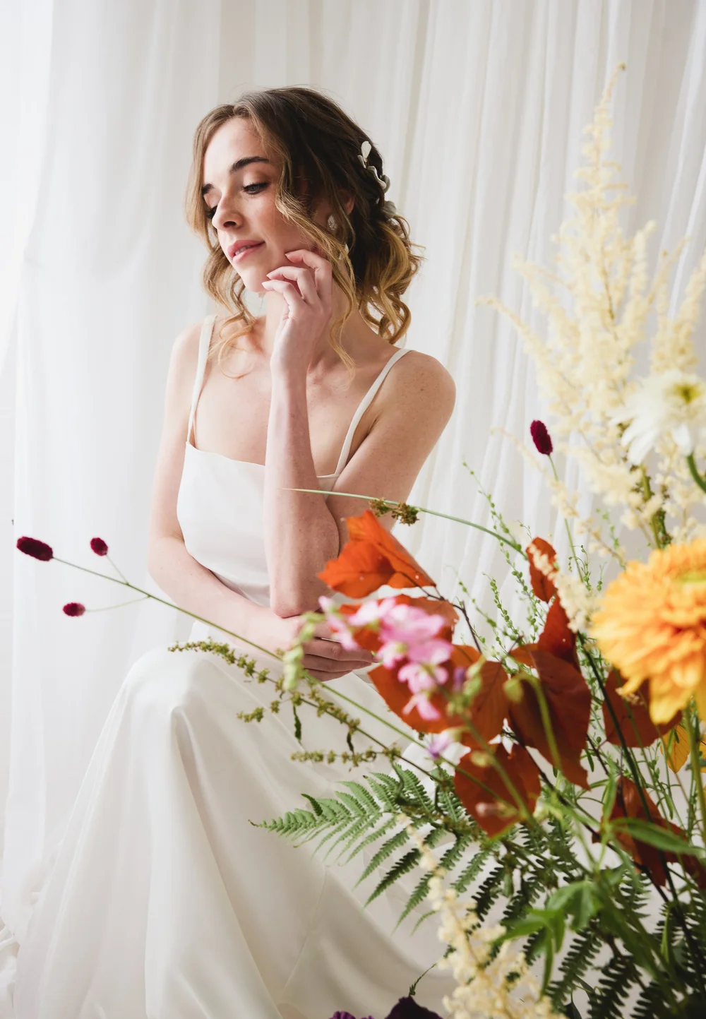 A young woman with wavy brown hair in a white dress, sitting in front of a white curtain, surrounded by colorful flowers.