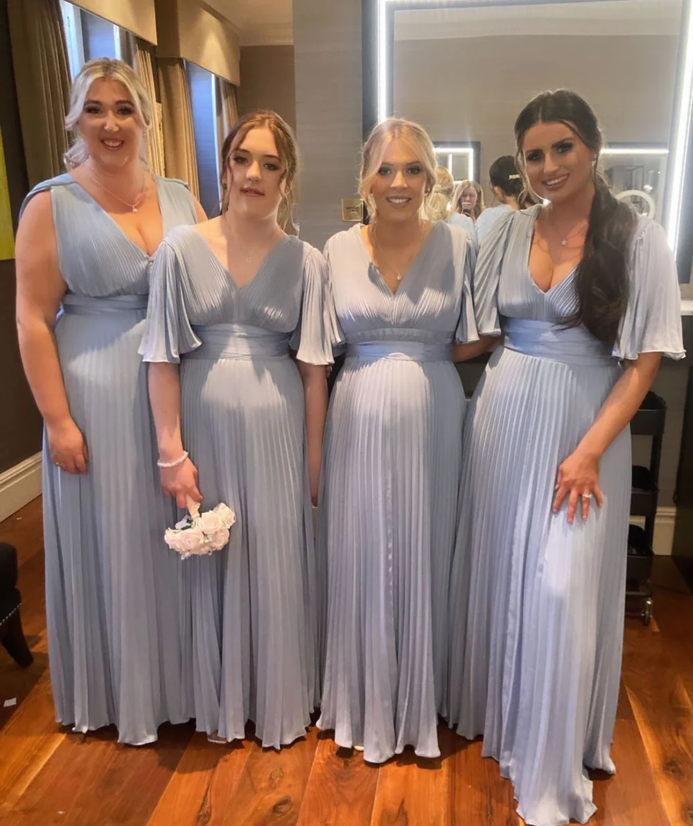 Four women in matching light blue pleated dresses standing in a room with wooden floors and a mirror in the background.