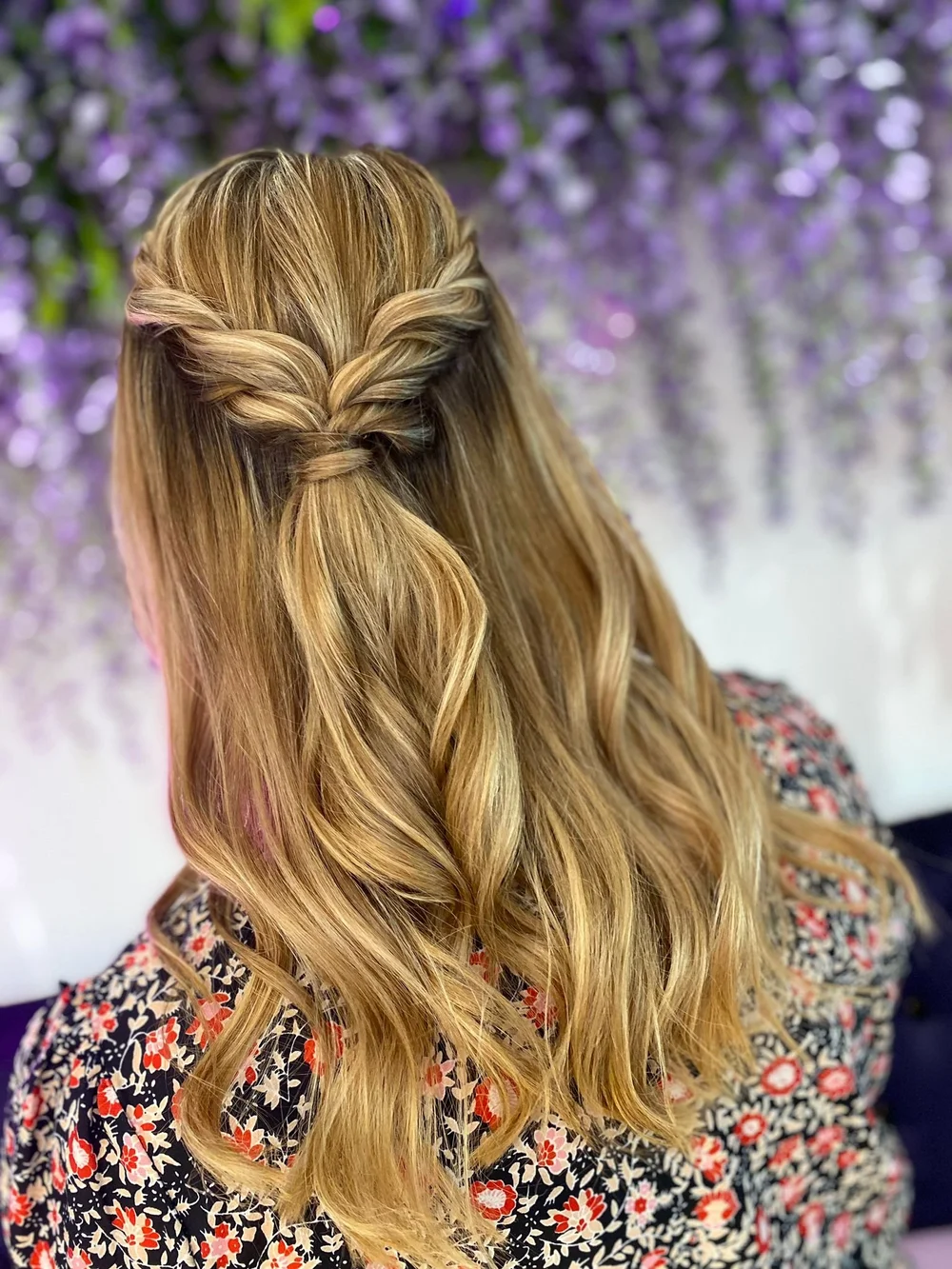 Woman with styled blonde hair in a half-up, half-down hairstyle with twisted sections, standing against a background of purple flowers.