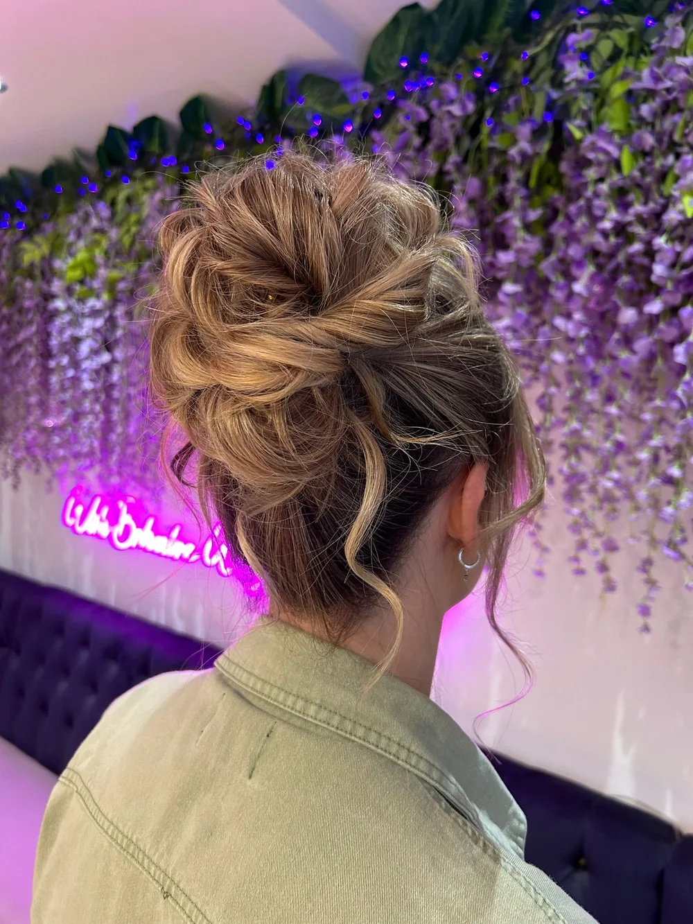 A woman with a curly updo hairstyle, wearing a light-colored jacket and earrings, is facing a wall decorated with purple flowers and purple string lights, with a pink neon sign in the background.