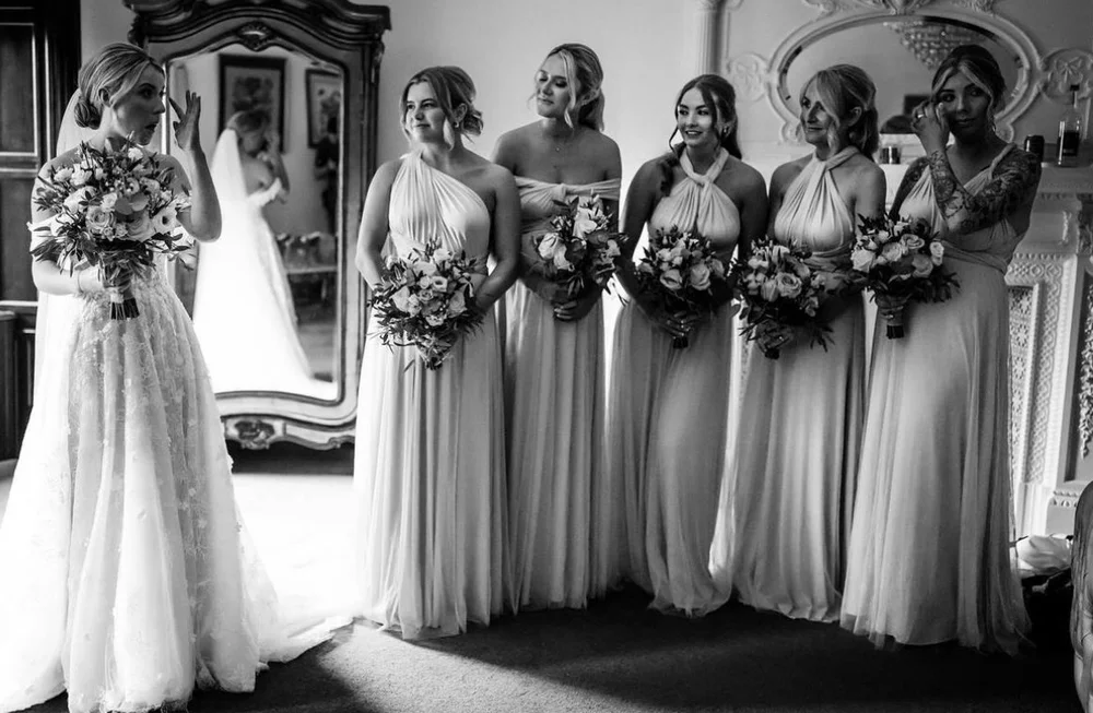 Six women in bridesmaid dresses holding bouquets, standing in a room with vintage furniture, with another woman in a wedding gown in the background, preparing for a wedding.