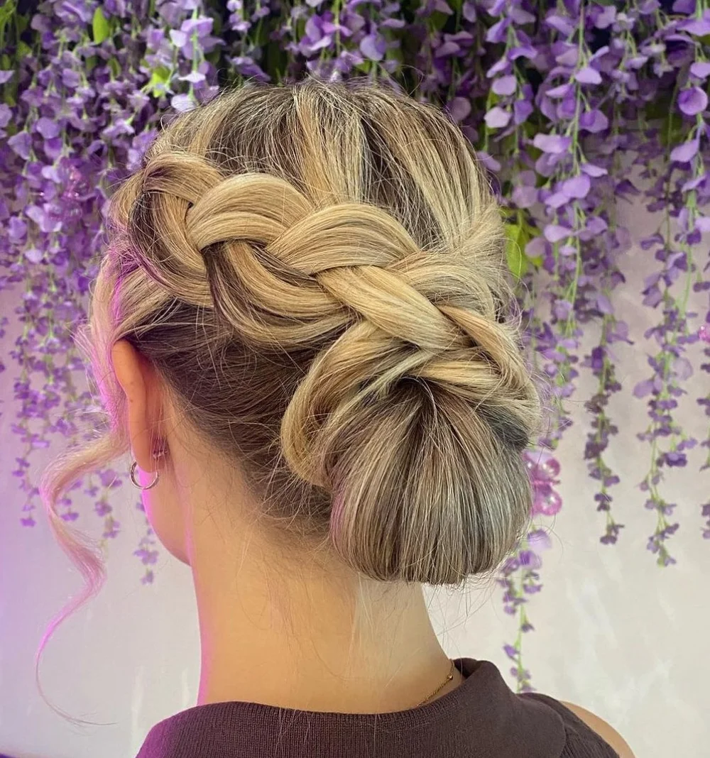 A woman with blonde hair styled in an elegant braided updo, with a loose curly strand on her left side, against a background of purple hanging flowers.