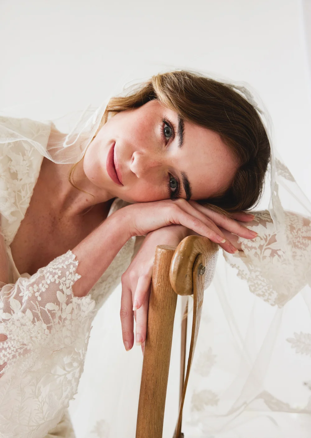 A woman with brown hair and blue eyes, wearing a lace dress, resting her head on her arm, leaning on a wooden chair with a floral background.
