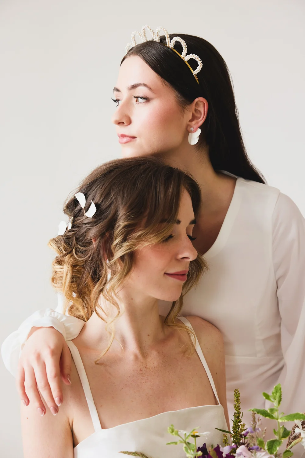 Two women with closed eyes, one wearing a pearl tiara and the other with white hair clips, embracing in a pose with a bouquet of flowers in front of them, against a plain white background.