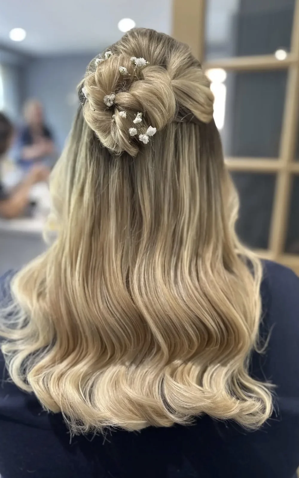 Back view of a woman with long, wavy blonde hair styled in a half-up, half-down hairstyle with a twisted bun and small white floral hair accessories.