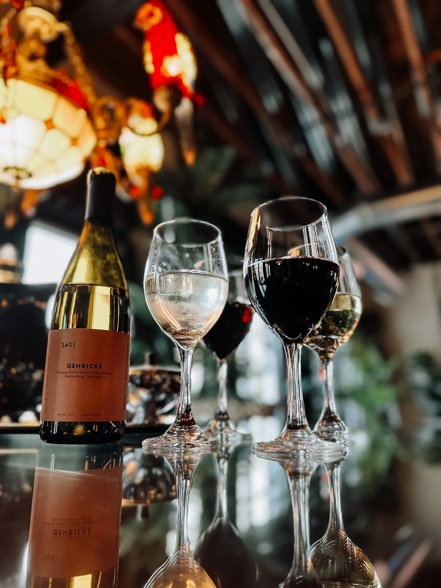 A wine bottle with a beige label and four wine glasses filled with white and red wine on a reflective surface in a dimly lit restaurant or bar with hanging decorative lamps in the background.