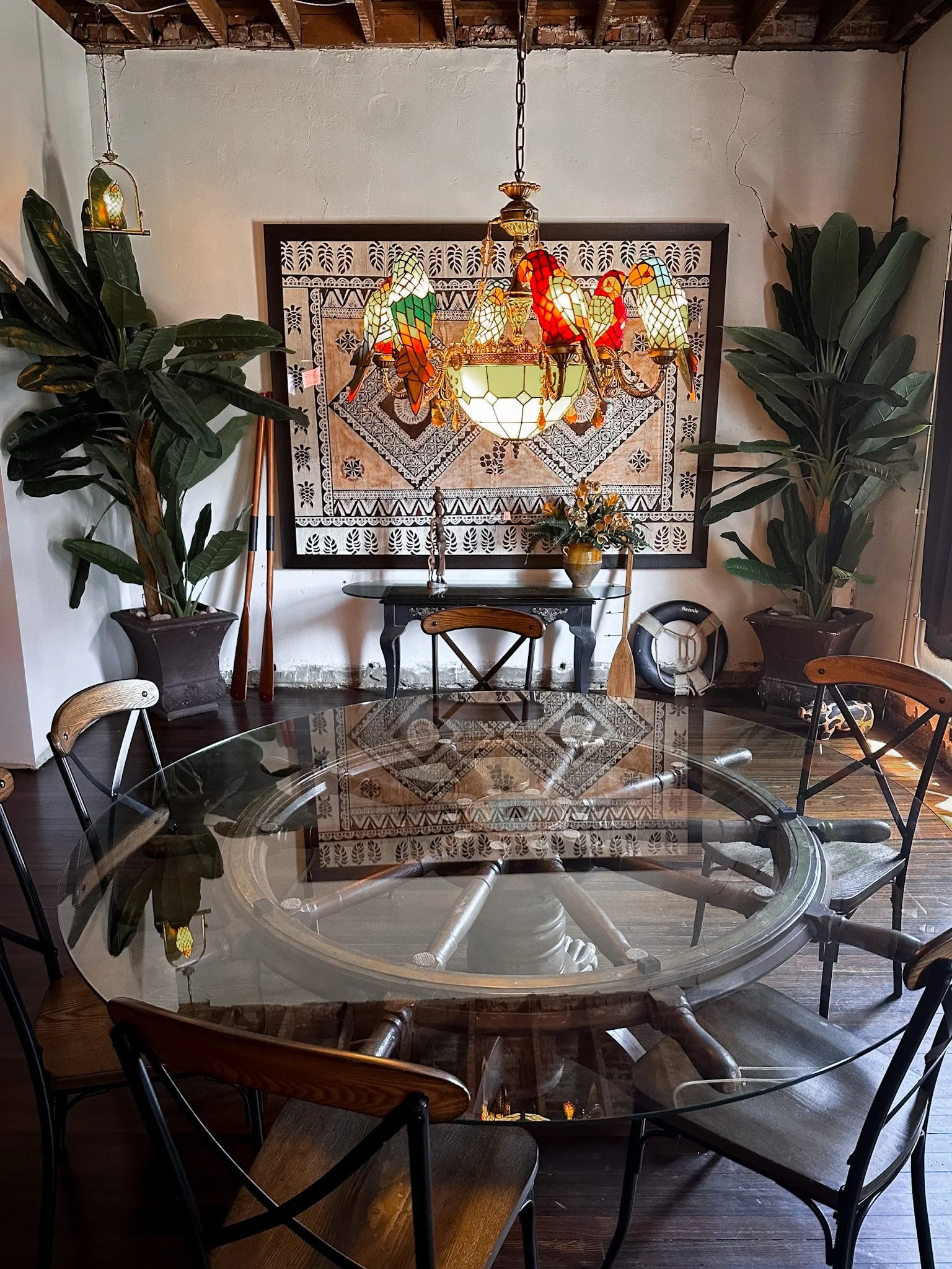 A room with a round glass-top dining table surrounded by wooden chairs, two large potted plants on each side of the room, and a chandelier with stained glass parrot designs hanging from the ceiling. The back wall is decorated with a large artwork fea