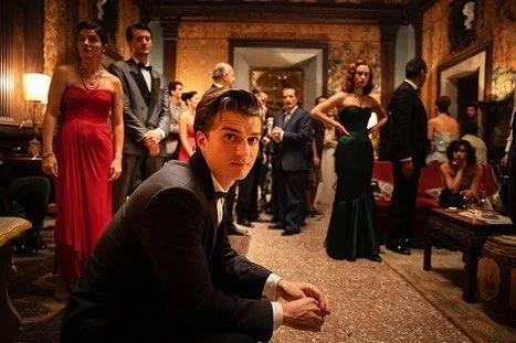 Young man in tuxedo sitting in a crowded, elegant room with other formally dressed guests engaging in conversations.