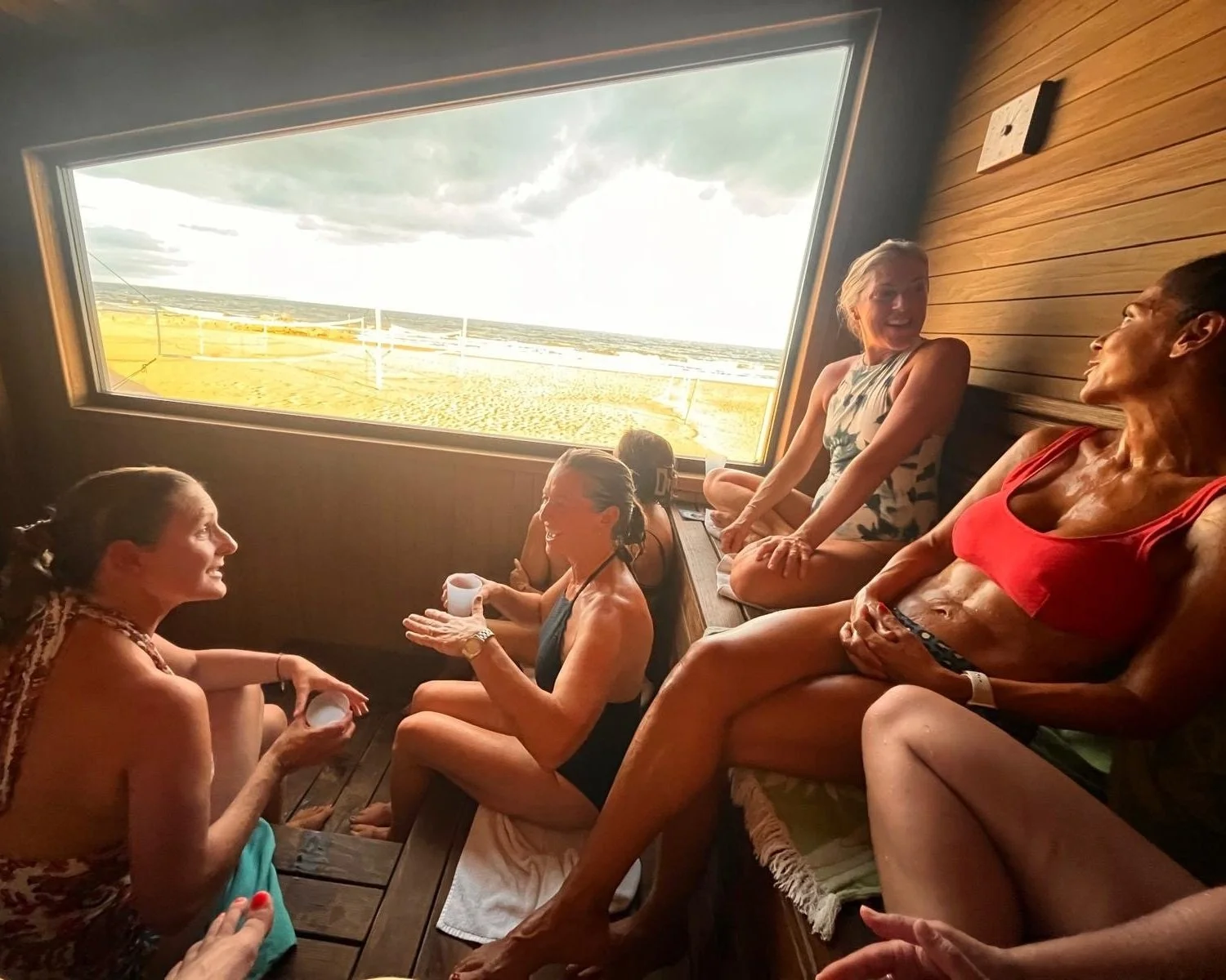 Five women enjoying a sauna with a large window overlooking a beach and cloudy sky.