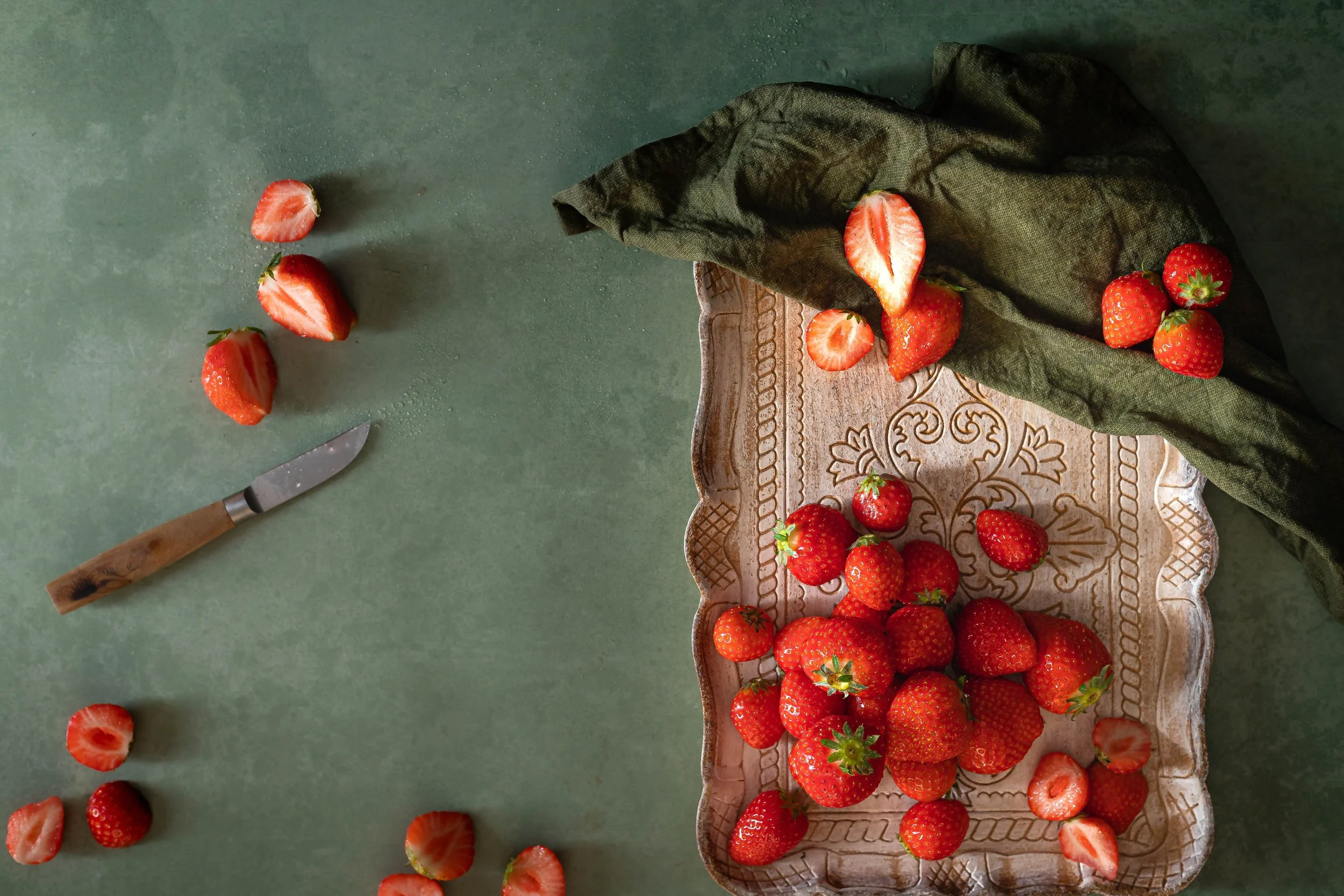 A rustic ceramic plate with strawberries on a green surface, some strawberries on a dark cloth, strawberry slices, and a small knife with a wooden handle.