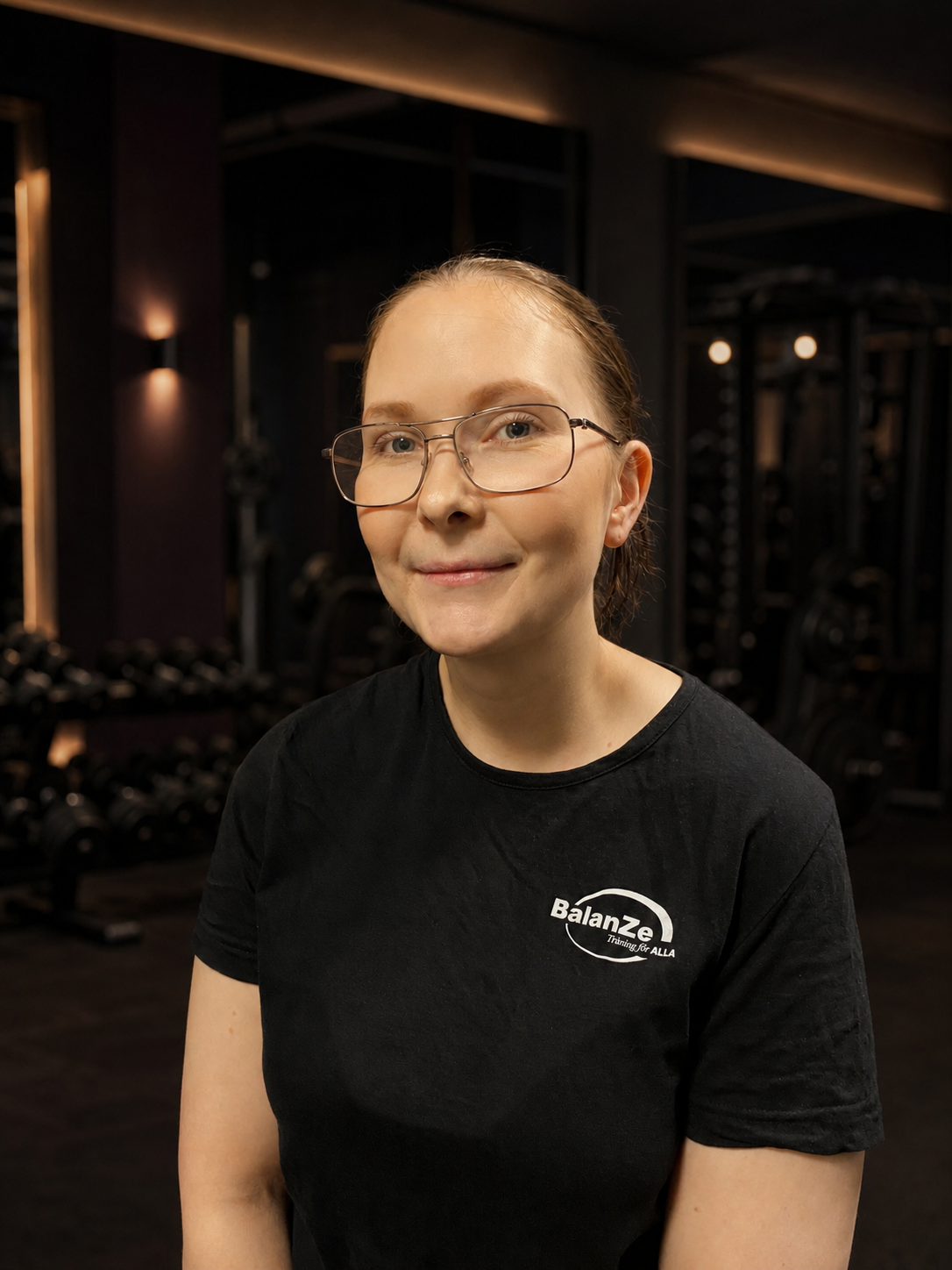 En person med glasögon och svart t-shirt står framför en mörk vägg.