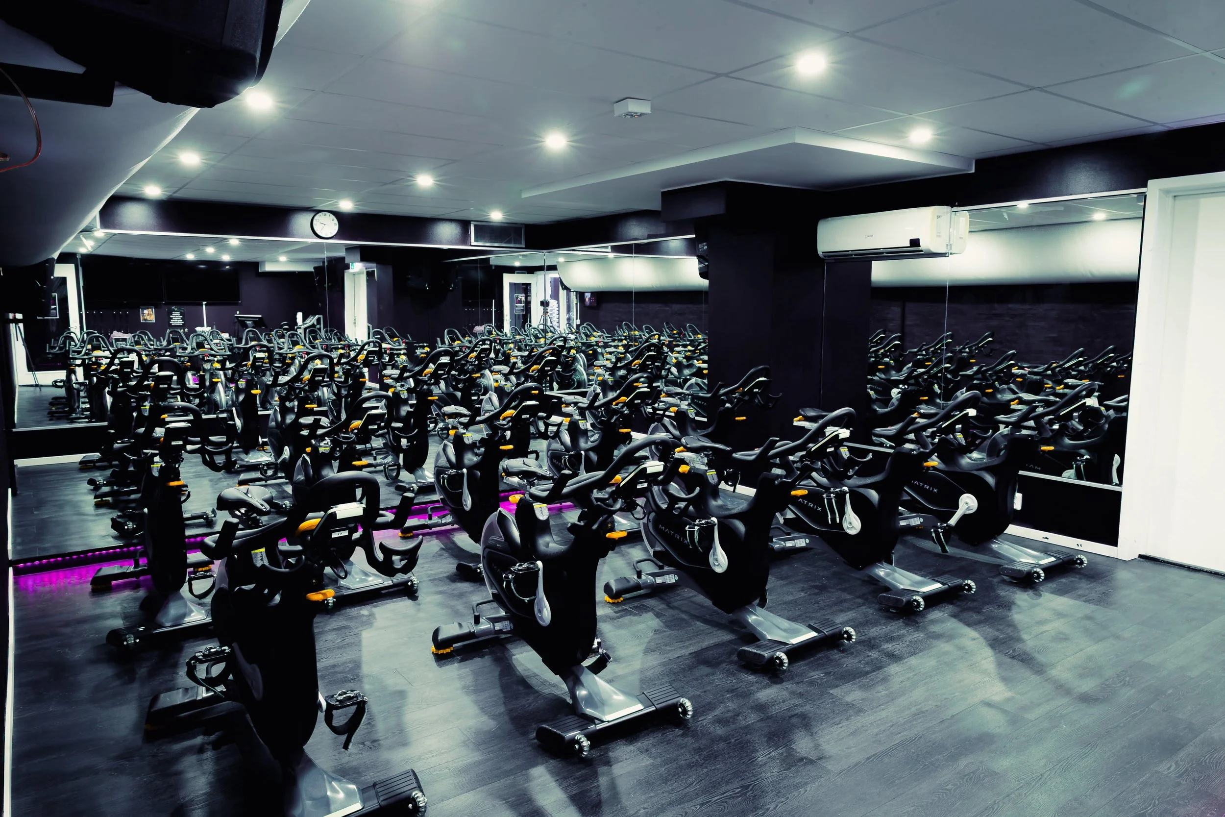 Ett mörkt rum med flera motionscyklar uppställda i rader, upplysta av lila ljus. Speglar finns på väggarna och taklampor lyser upp lokalen.