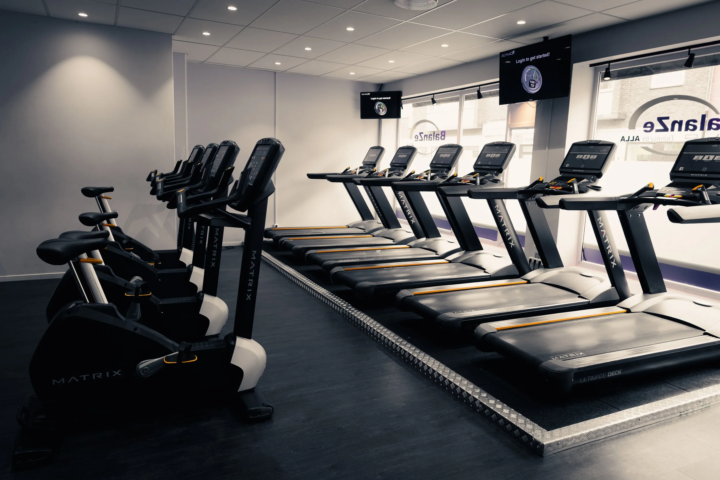 Interiör av ett gym med flera löpband på rad och träningsmaskiner. Ljusa väggar och stort fönster.