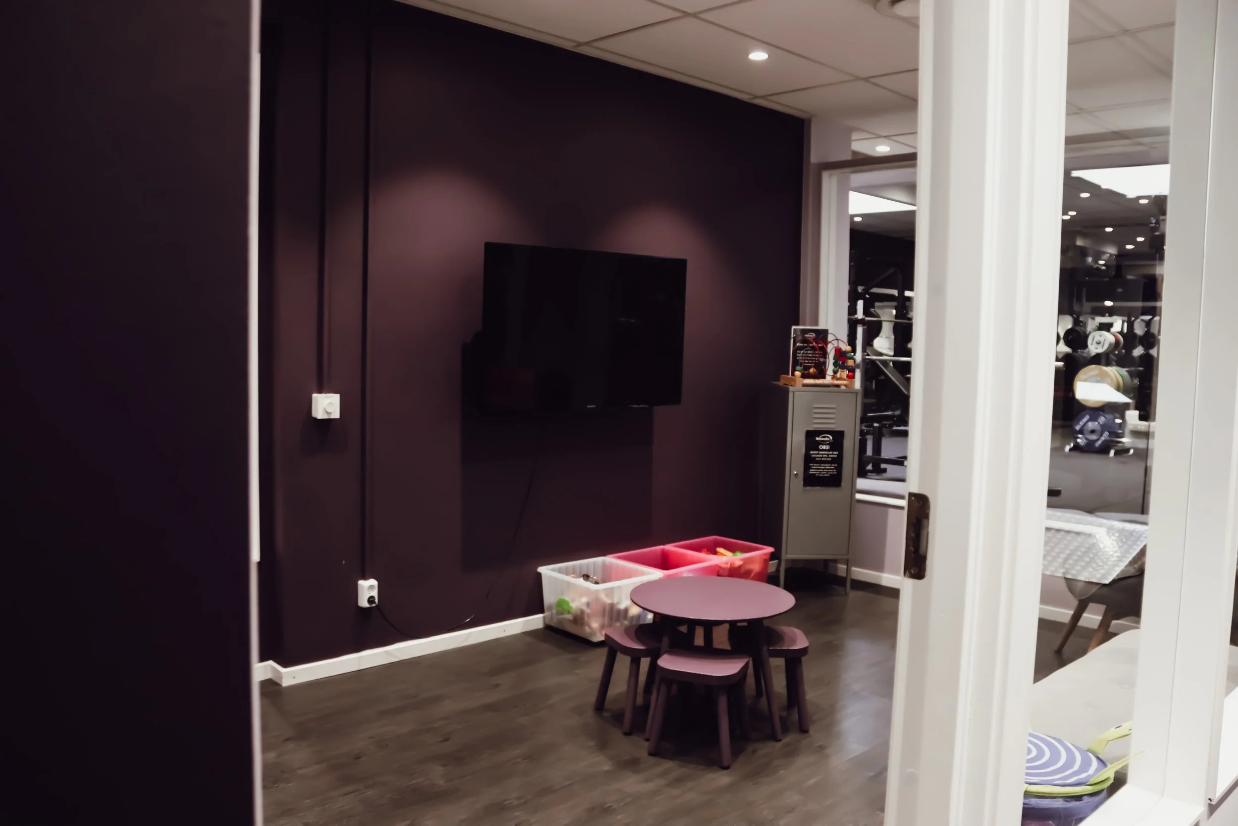 Litet inomhusområde med ett runt bord och stolar för barn, en väggmonterad TV, färgglada lådor och ett gym synligt genom fönstret.