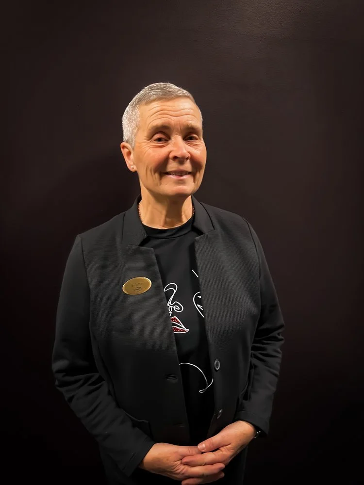 En äldre person med kort grått hår, klädd i en svart kavaj, står framför en mörk bakgrund och ler mot kameran.