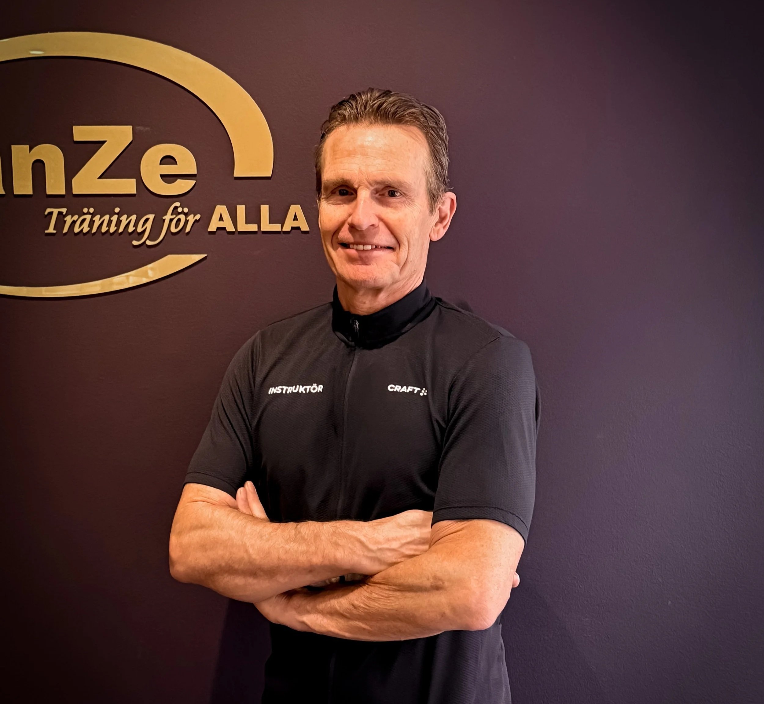 En person med lila t-shirt står mot en beige vägg. Texten på t-shirten lyder "Balanze Träning för alla."