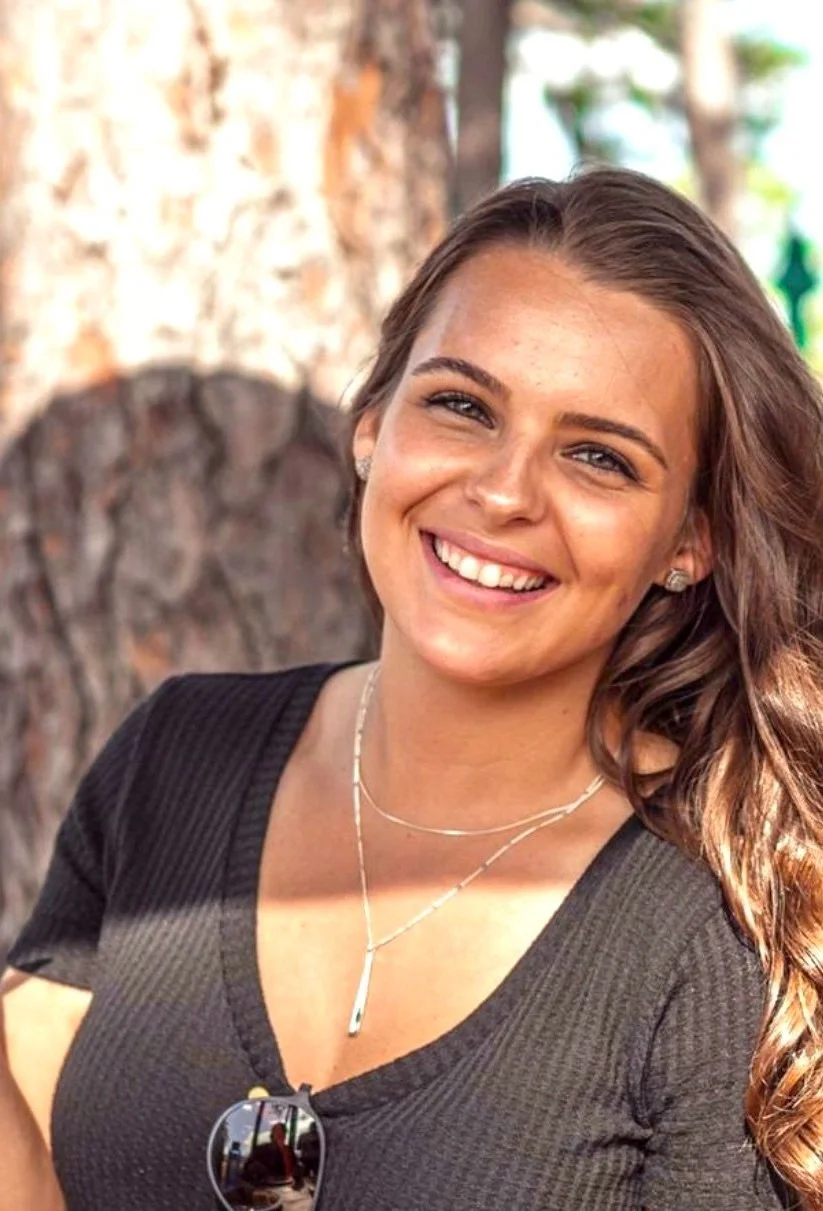 A smiling woman with wavy brown hair wearing a black shirt, layered necklaces, and earrings, standing outdoors with trees in the background.