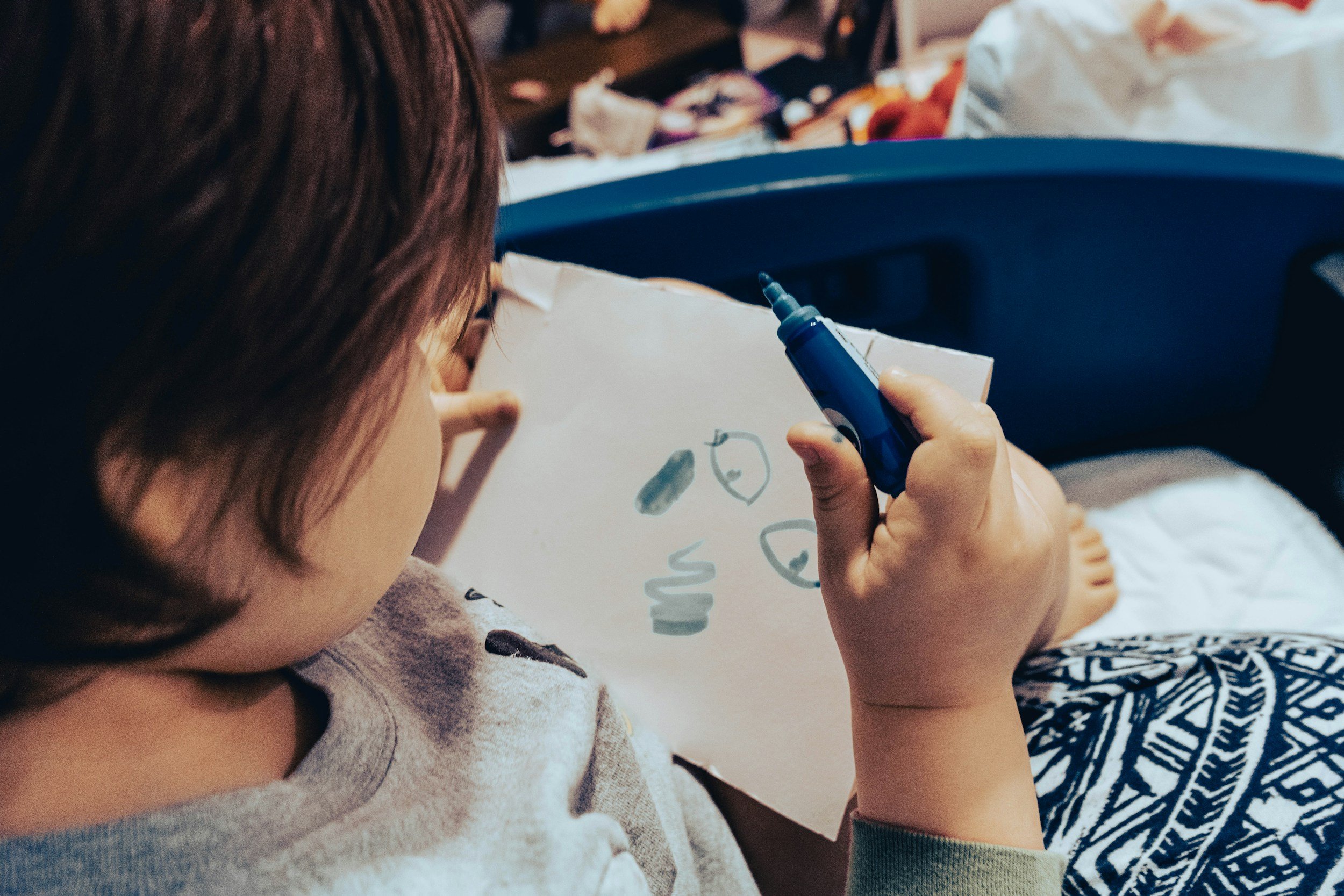 A young child with brown hair and glasses drawing on a white piece of paper with a blue marker. The drawing includes simple shapes and faces. The child is sitting in a blue chair in a cluttered room.