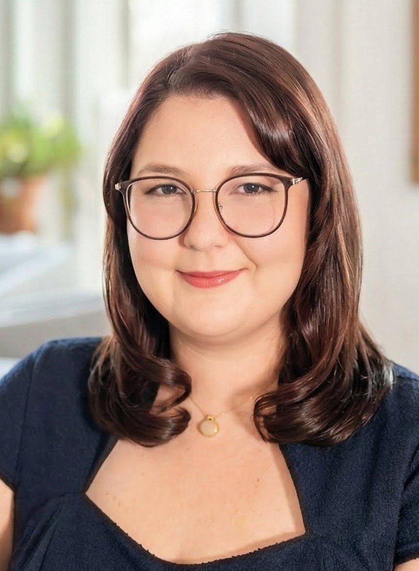 Close-up of a woman with long dark wavy hair, light skin, and a slight smile, wearing a black shirt, standing indoors against a neutral background.
