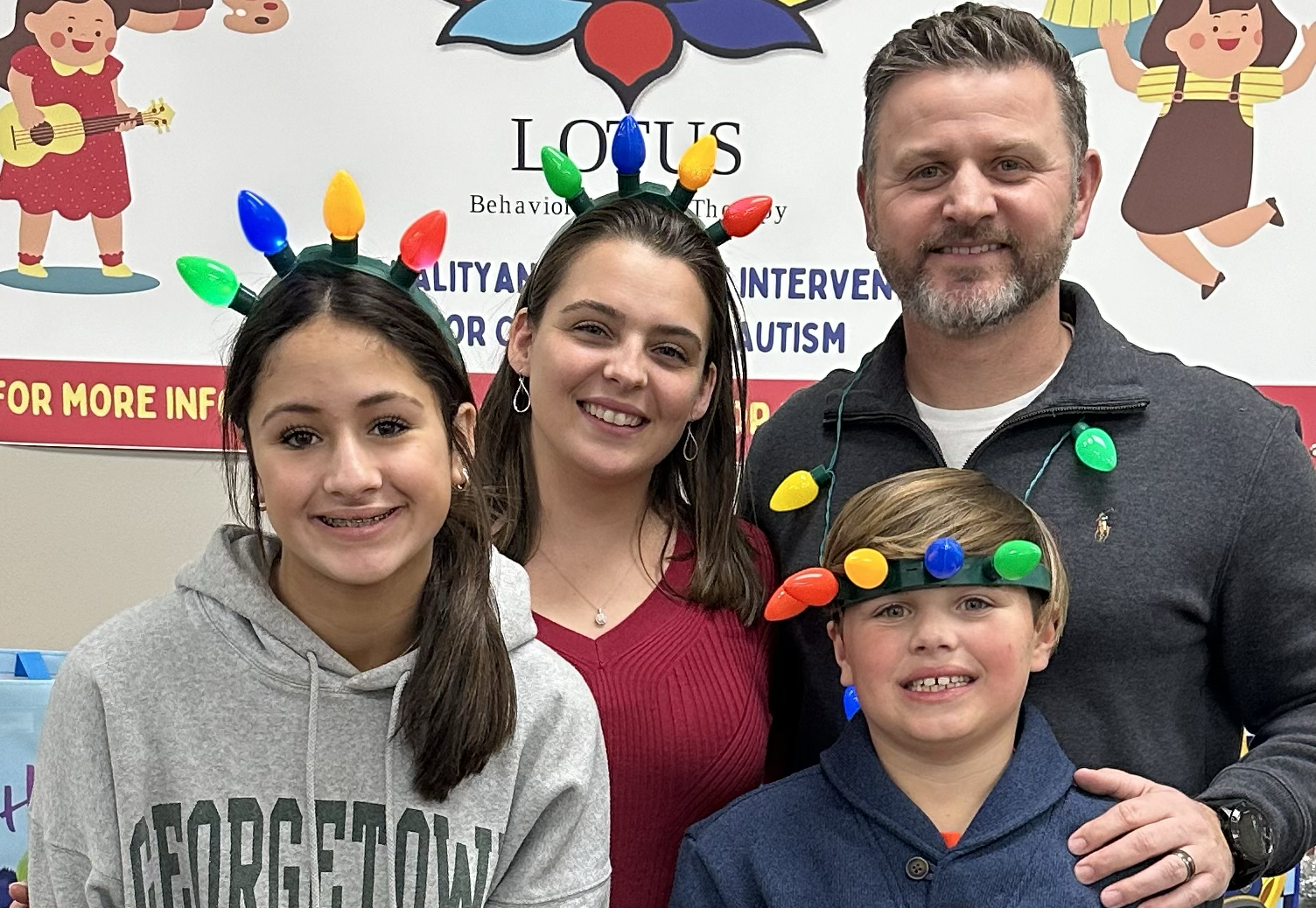 The owners, Michael and Brittany, and their two kids, are smiling at a holiday event. They are wearing festive light-up headbands with colorful lights. Behind them is a poster about autism intervention and therapy.