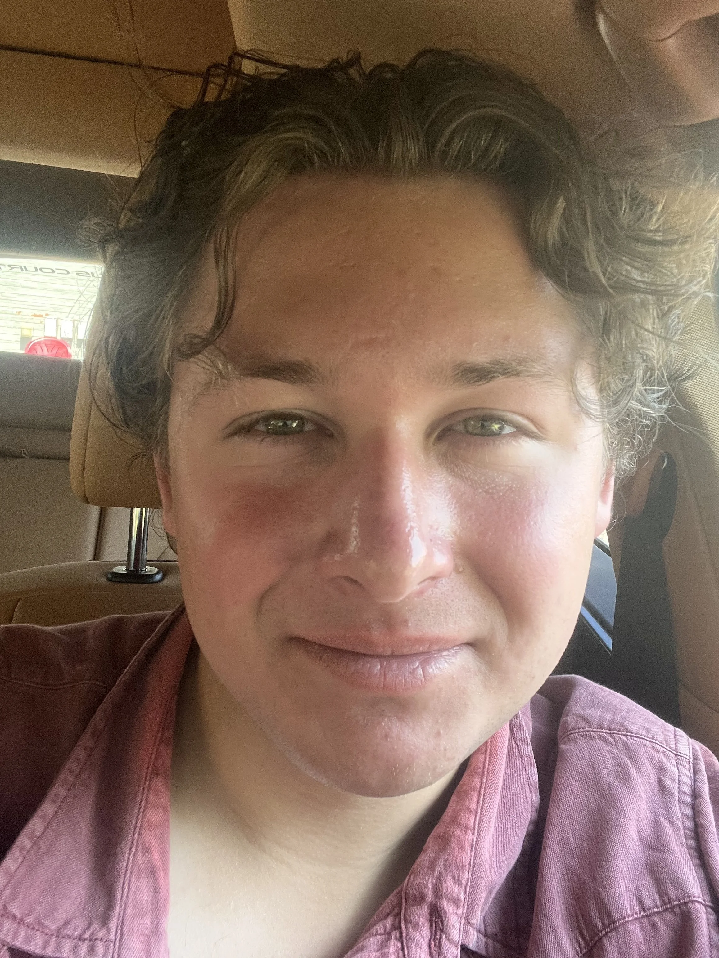 Close-up of a young man with light skin, curly brown hair, and a slight smile, sitting inside a vehicle with a beige interior.