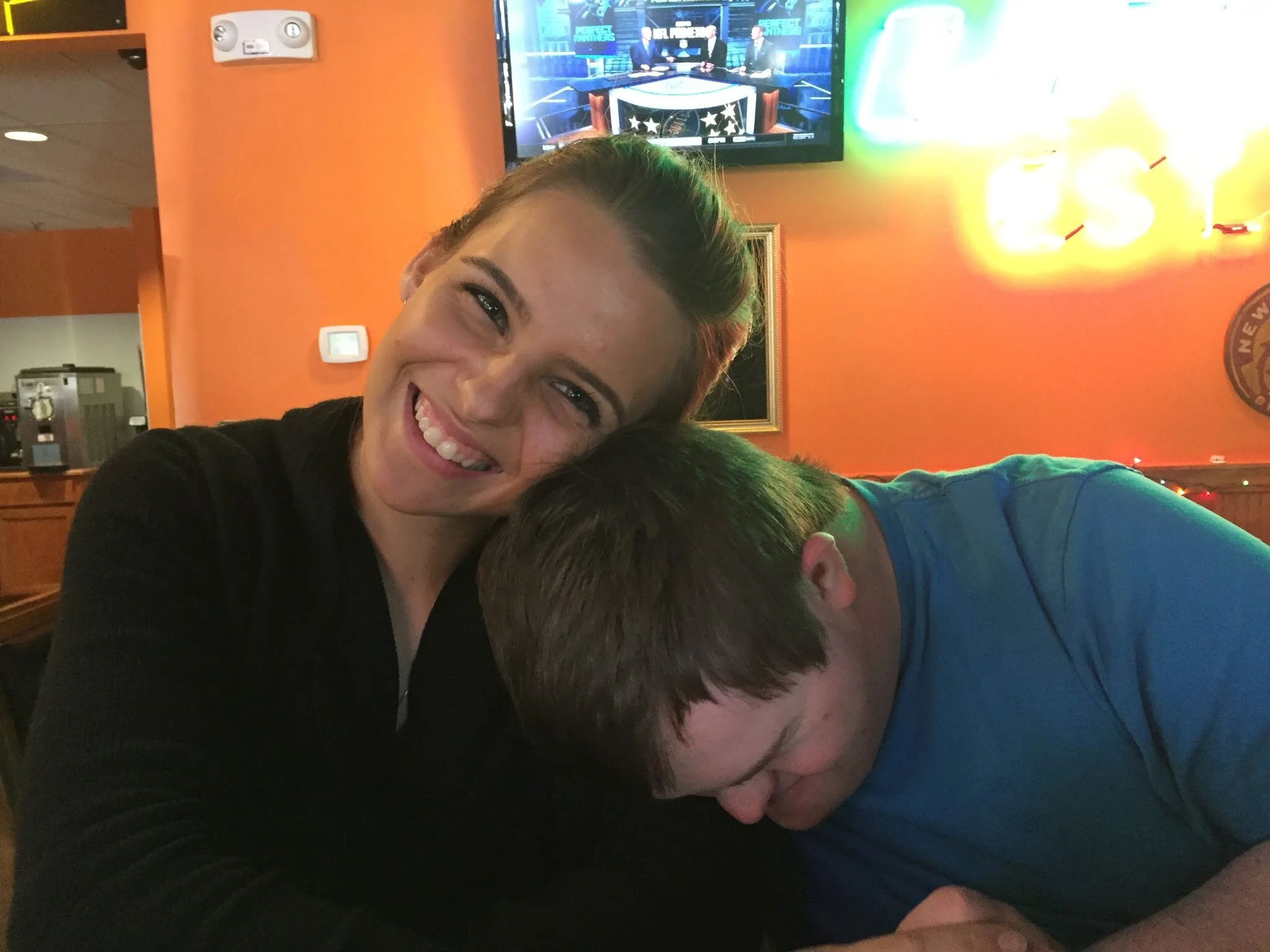 Brittany Evans, smiling, leaning in with a client, inside a restaurant or bar with neon lighting and a TV screen in the background.