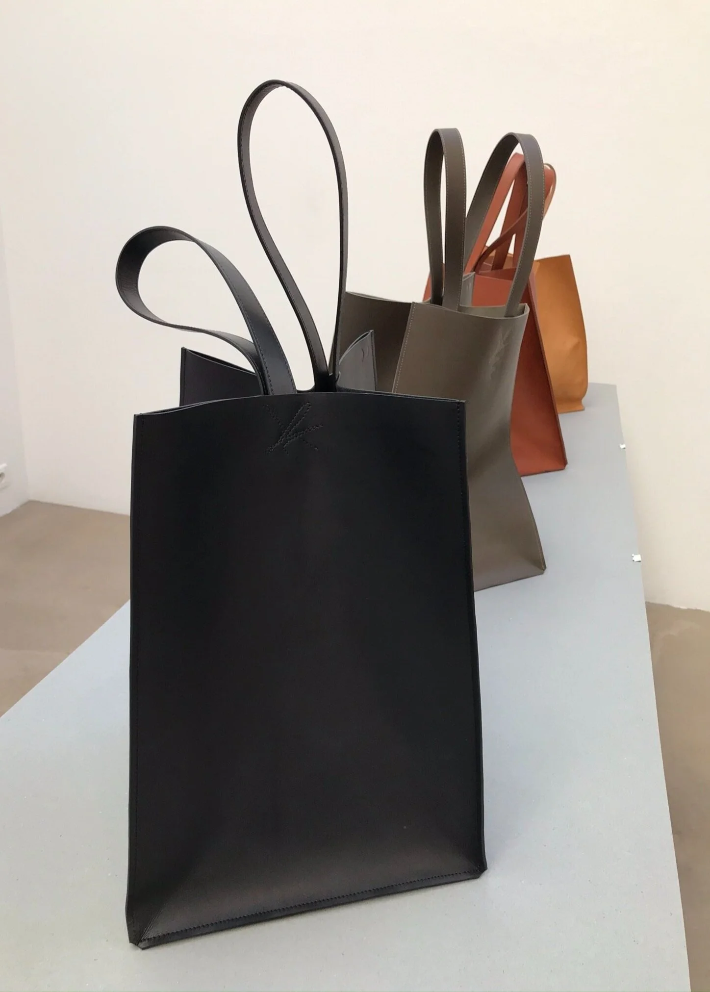 Three minimalist leather tote bags in black, gray, and tan colors, arranged on a white table against a plain white wall.