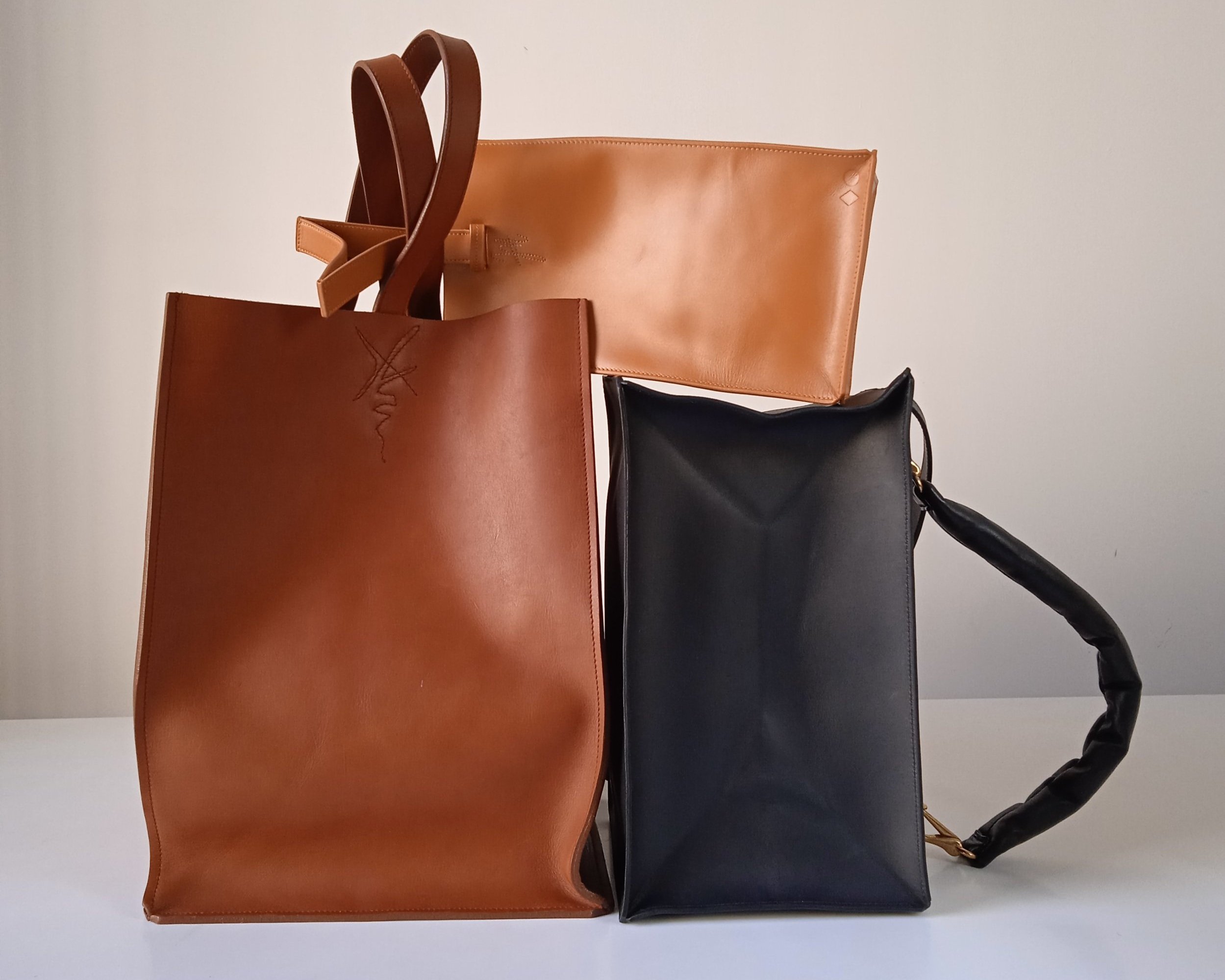 Three leather bags in brown, tan, and black, arranged on a white surface against a plain wall.